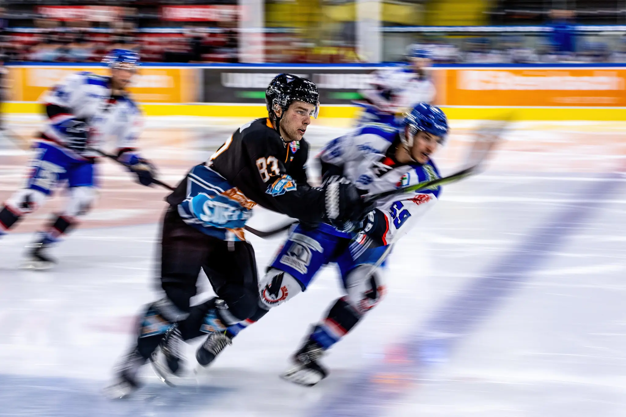 Ein Eishockeyspieler der SteelWings Linz im schwarzen Heimtrikot und ein gegnerischer Spieler in blau-weißem Trikot sind in schneller Aktion auf dem Eis zu sehen