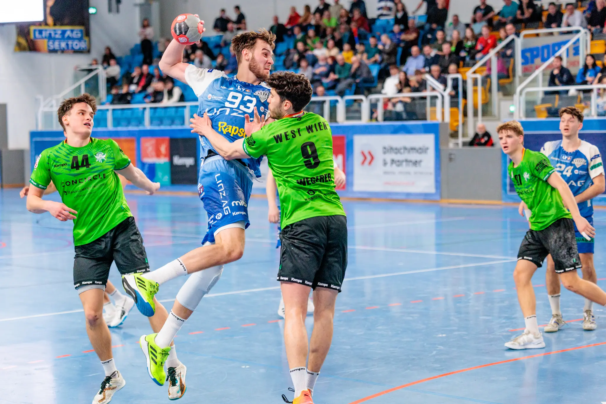 Dynamisches Handballspiel mit einem Spieler im blauen Trikot (Nr. 93) im Sprungwurf, verteidigt von Spielern in grünen Trikots, auf einem blauen Spielfeld 