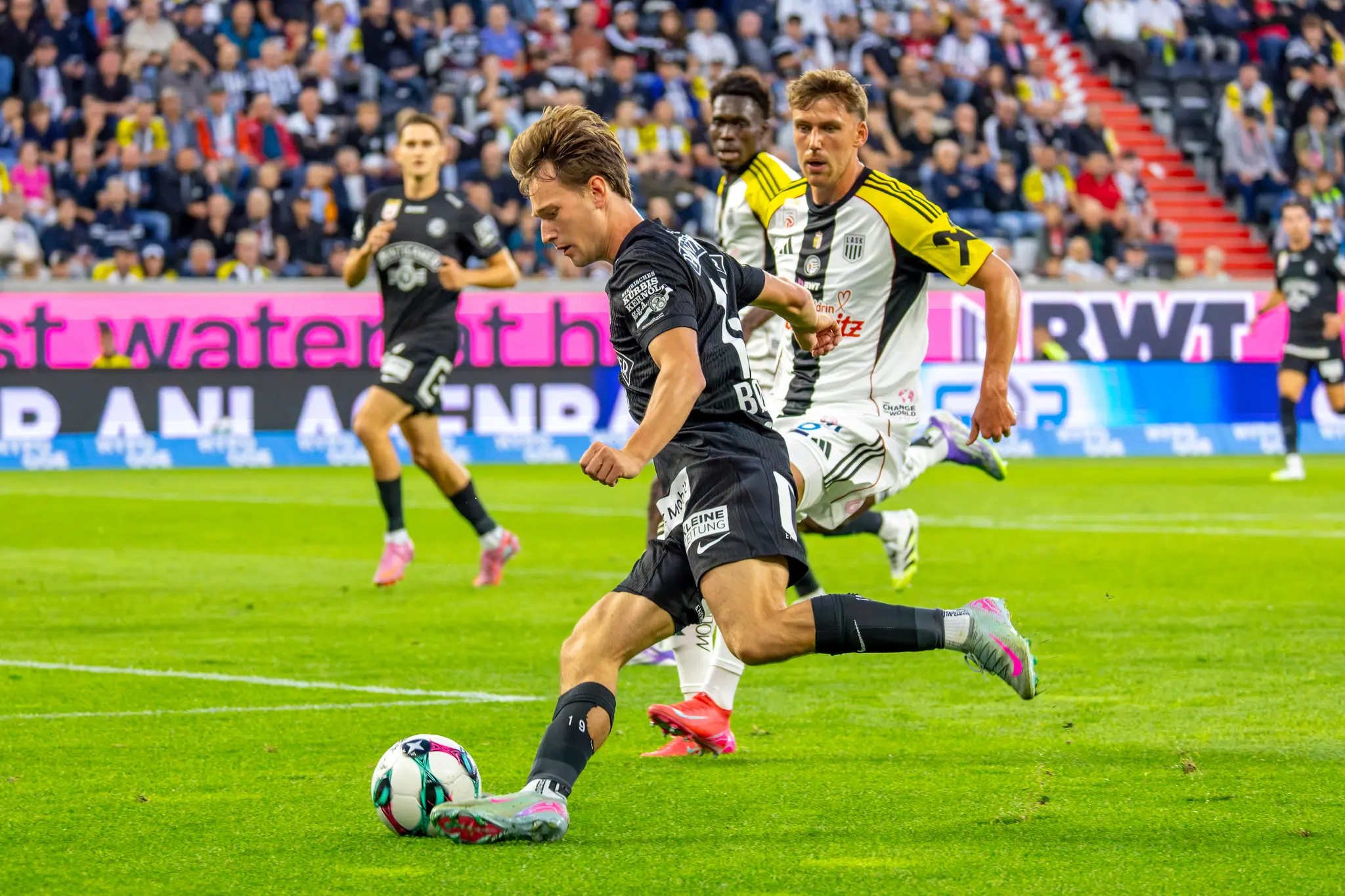 Ein Fußballspieler von Sturm Graz im schwarzen Trikot schießt den Ball während eines Bundesligaspiels, verfolgt von einem LASK-Spieler