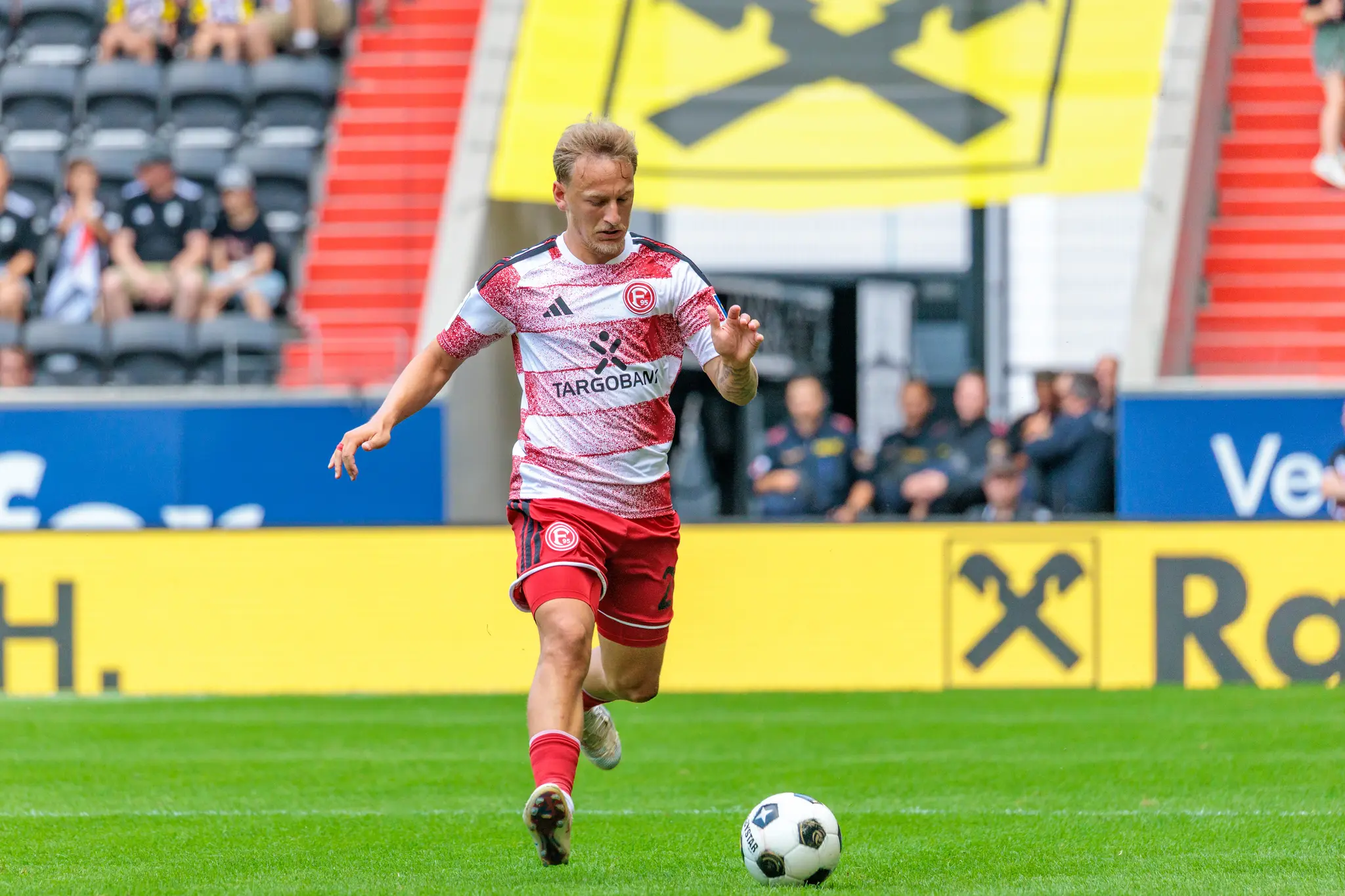 Fortuna-Düsseldorf-Spieler dribbelt den Ball, im Hintergrund das große gelbe LASK-Wappen an der Raiffeisen Arena.
