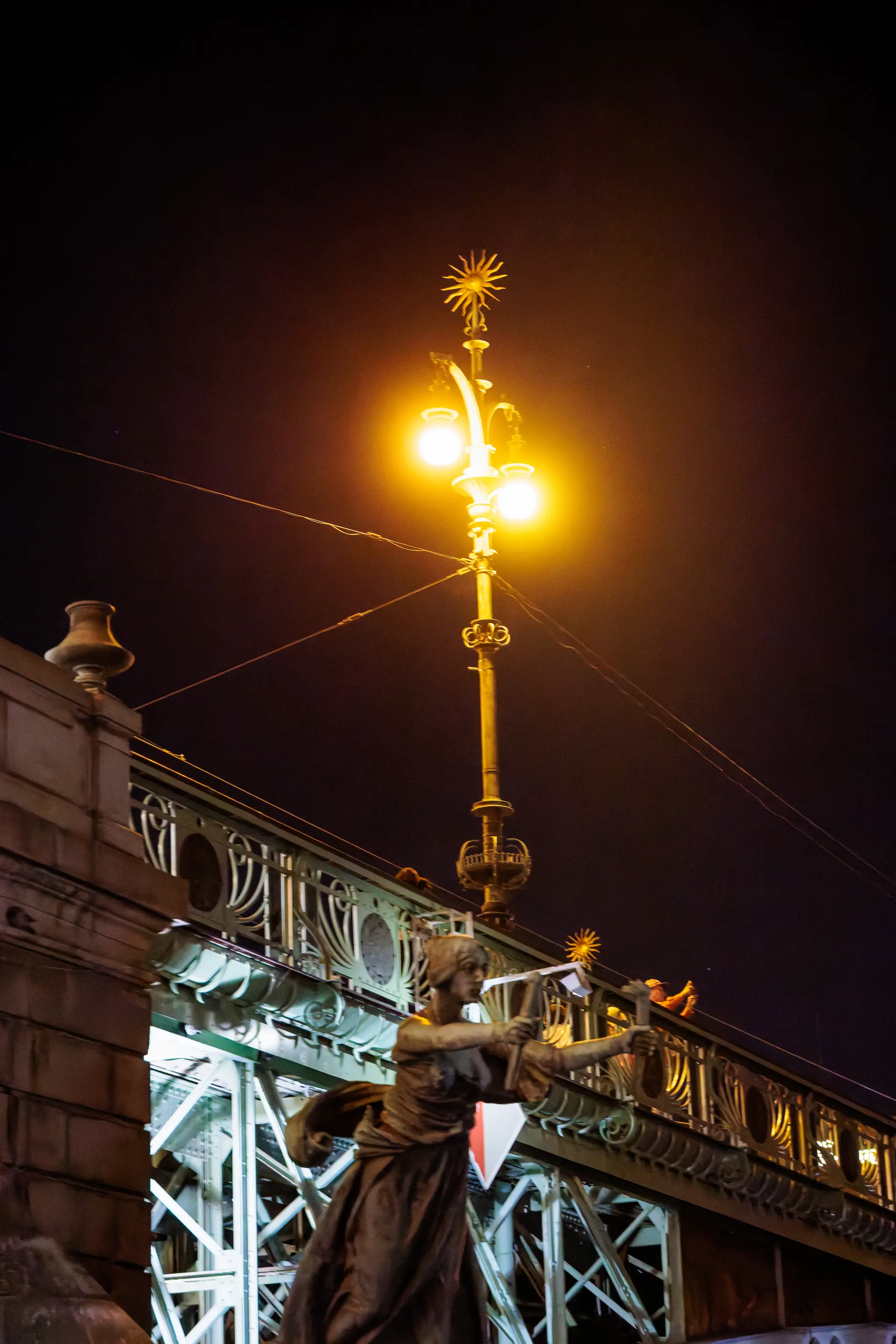 Bronzene Jugendstilfigur mit Hammer und Sternenlaterne an der Tschech-Brücke in Prag; Straßenbahn-Oberleitungen und warmes Laternenlicht prägen die Nacht.