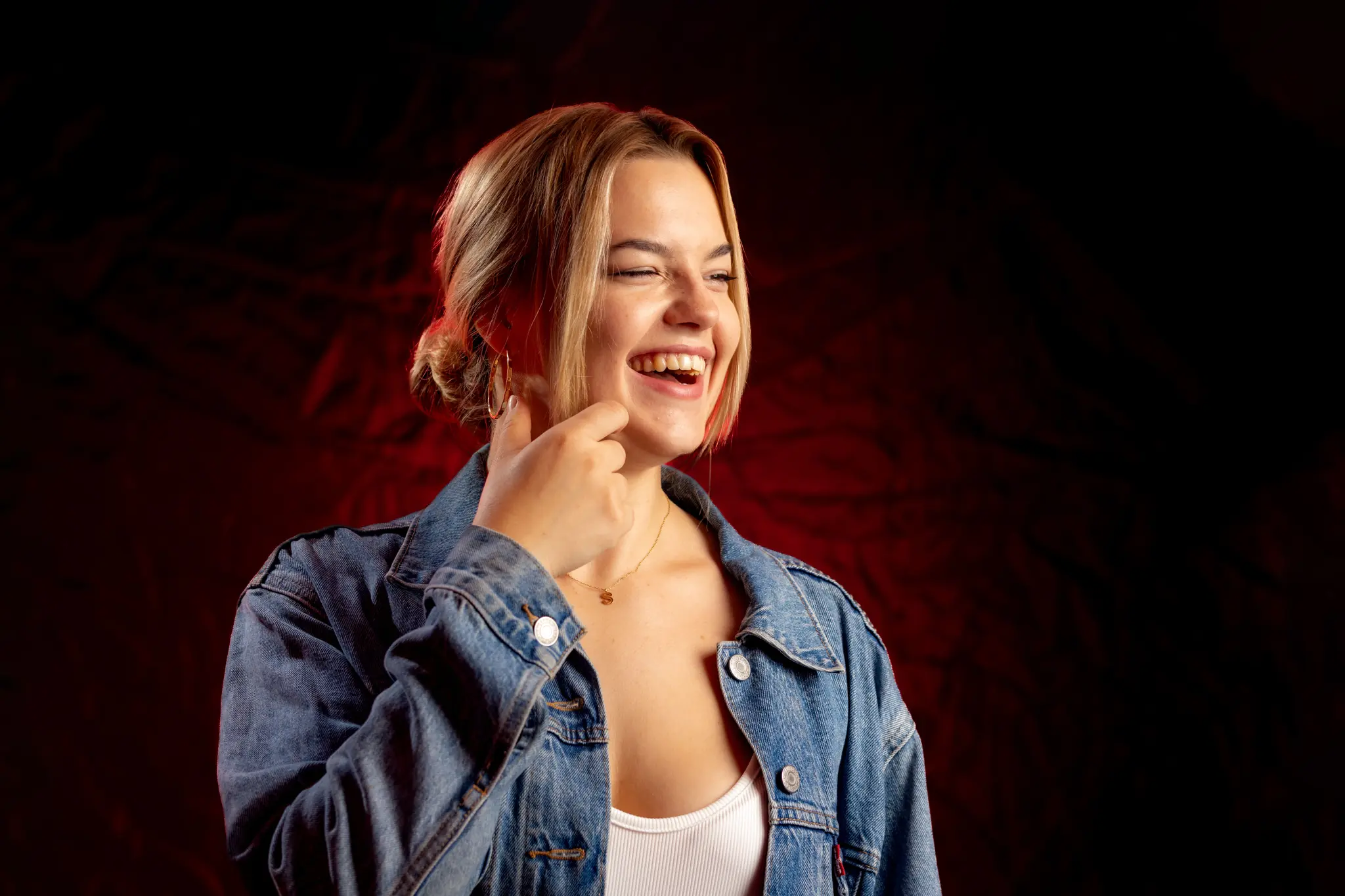 Eine junge lachende Frau mit blonden Haaren trägt eine Jeansjacke vor einem stimmungsvollen roten Hintergrund