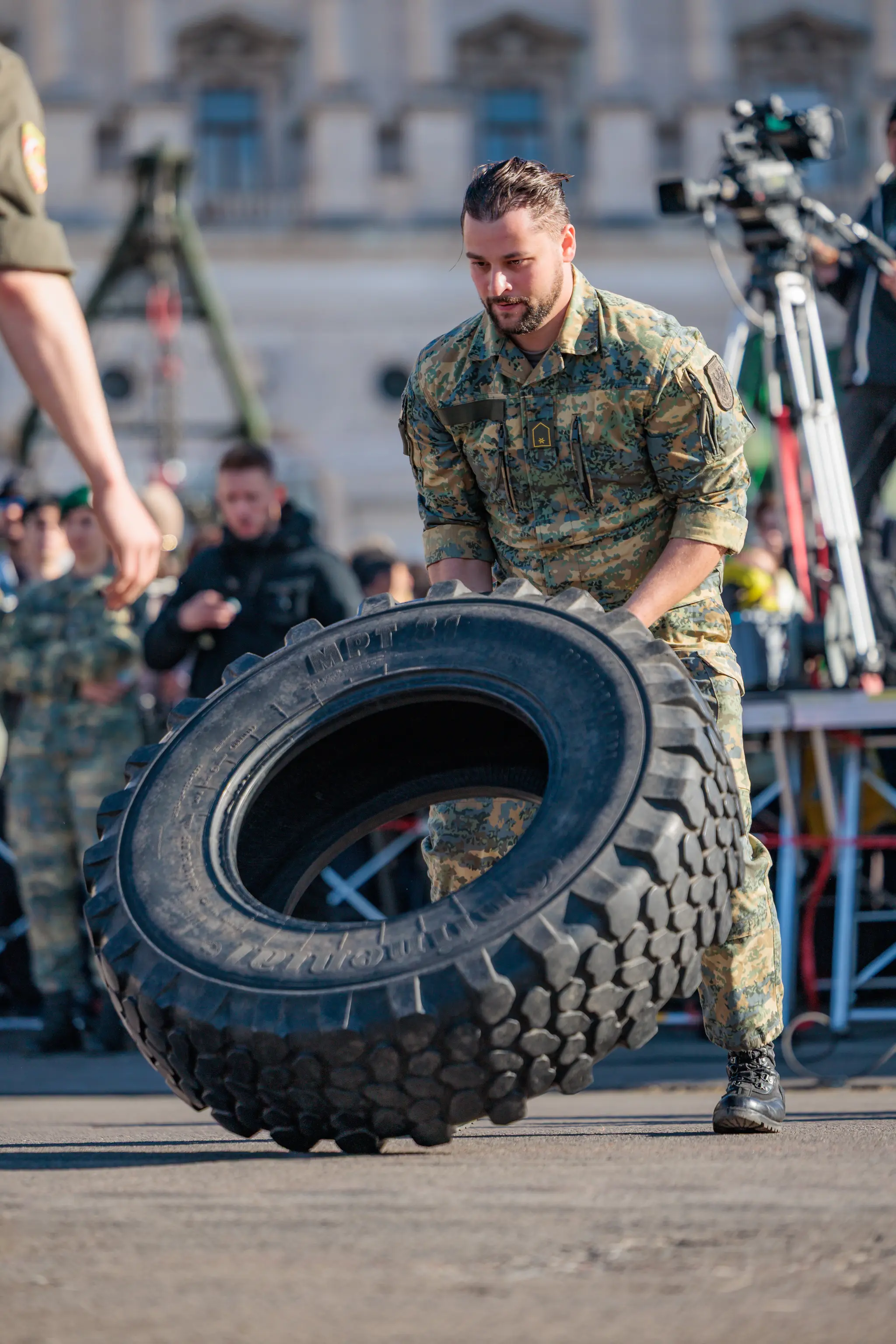Ein Soldat in Tarnuniform bewegt beim Heeressport einen großen Lkw-Reifen, im Hintergrund stehen Zuschauer und ein Kamerateam.