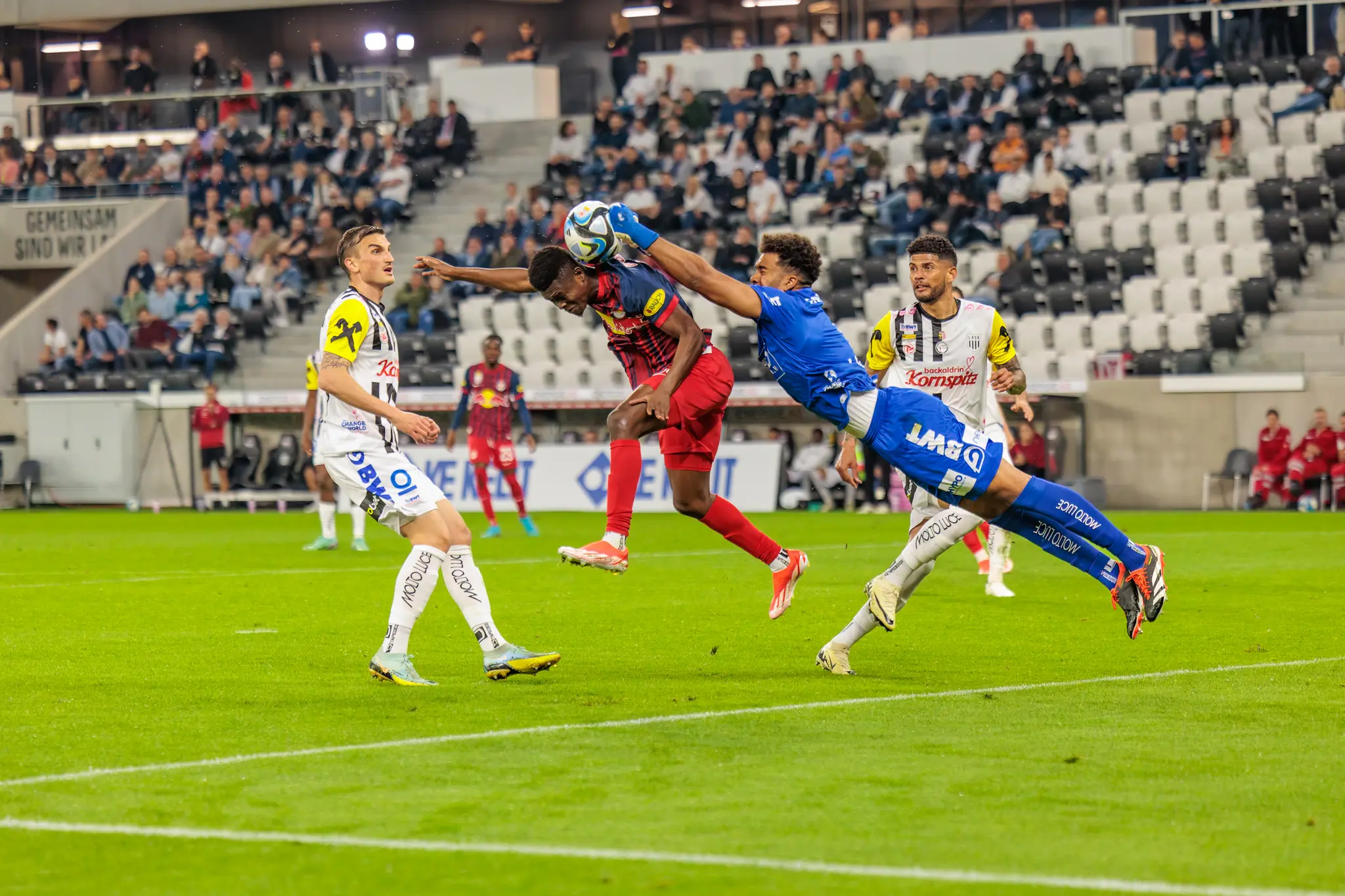 Ein dramatisches Kopfballduell zwischen dem Torwart im blauen Trikot und einem Spieler im roten Trikots während eines Fußballspiels in einem gut besuchten Stadion