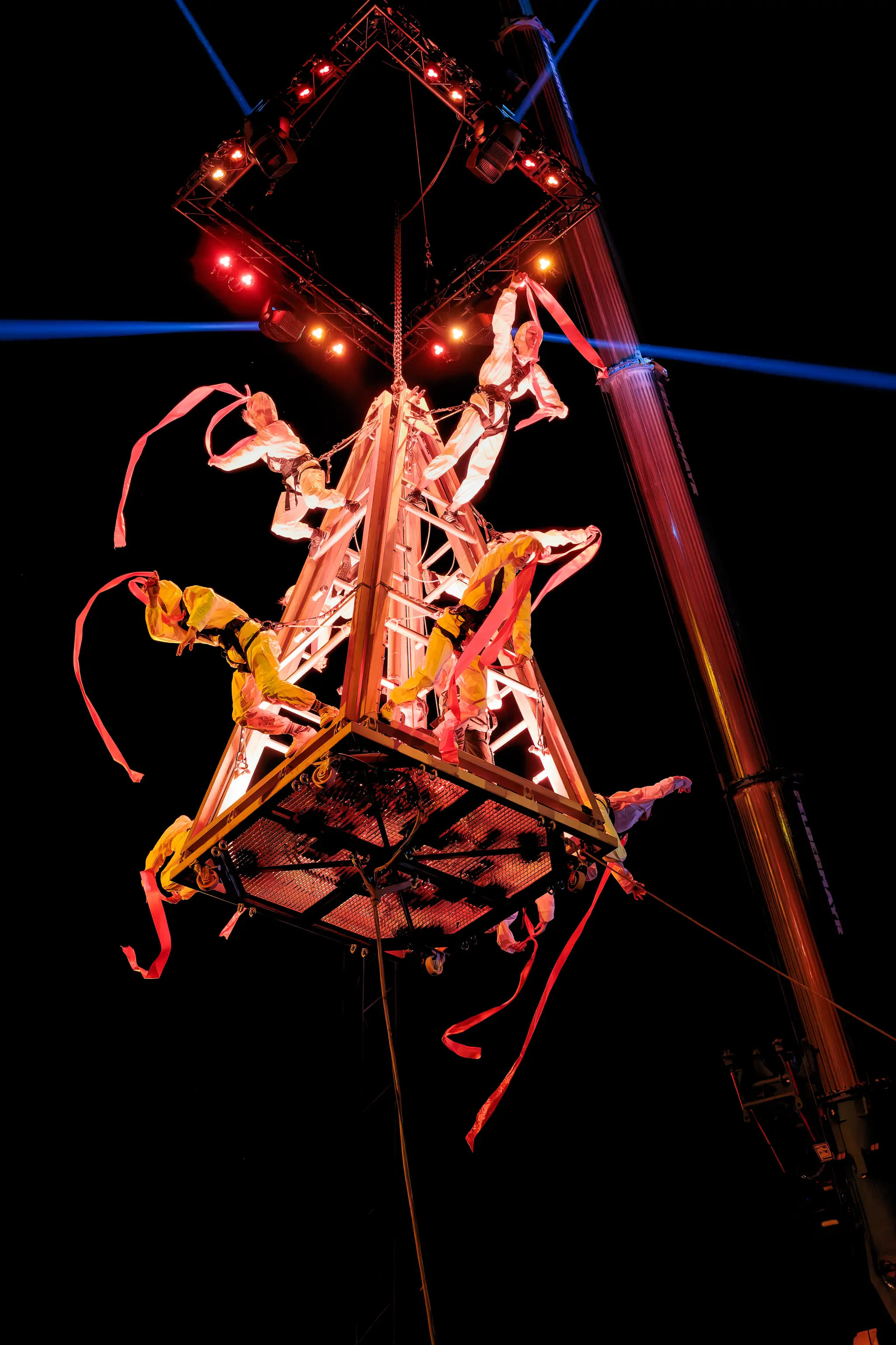 Spektakuläre Luftakrobatik-Show bei Nacht während der Linzer Klangwolke 2024: Mehrere Künstler in weißen und gelben Kostümen performen dynamisch an einer rot beleuchteten Hängekonstruktion mit roten Bändern. Blaue Lichtstrahlen im Hintergrund. 