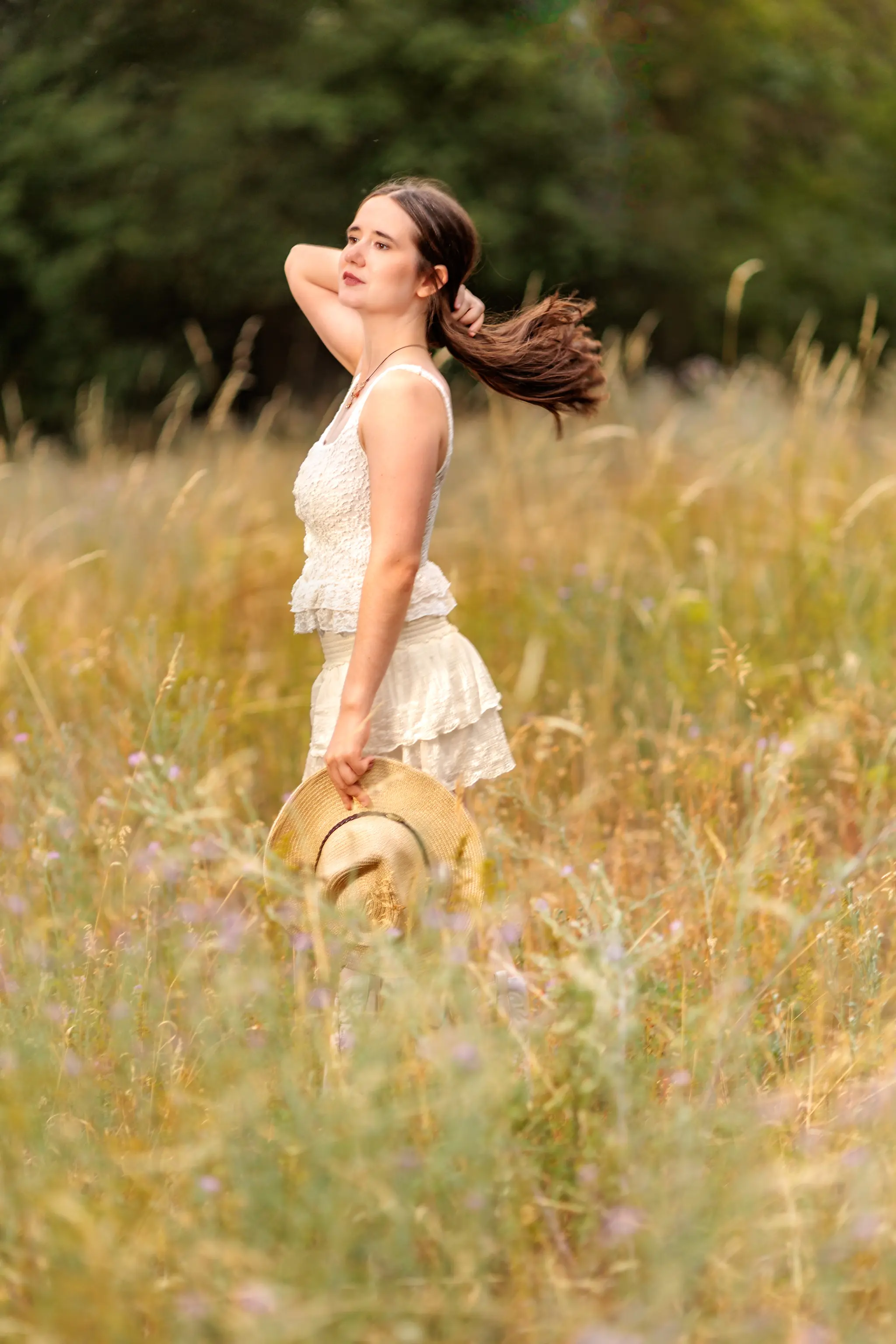 Hanna steht im hohen goldenen Gras einer Wiese, ihr Haar weht im Wind, während sie ihren Strohhut in der Hand hält.