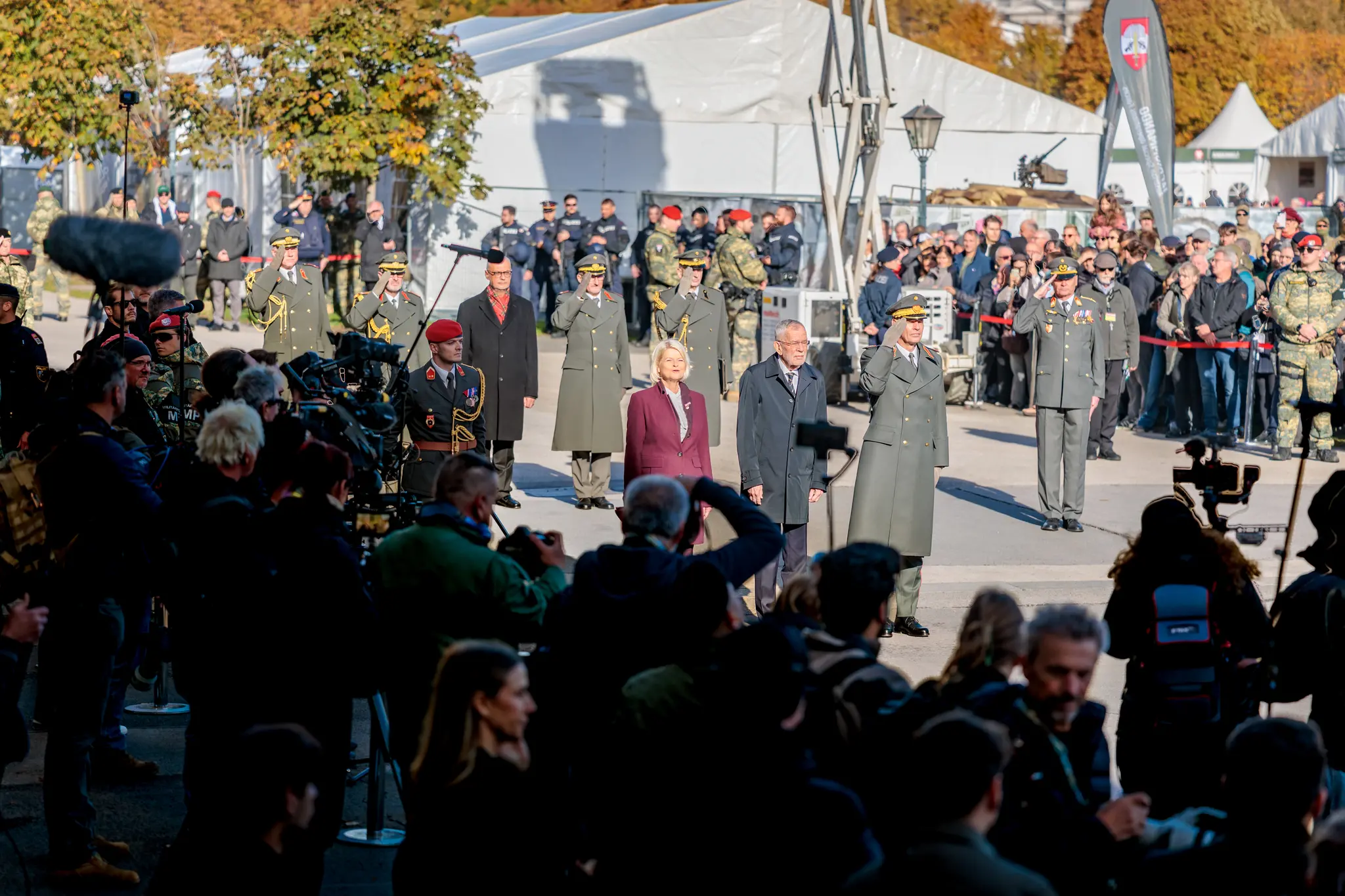Bundespräsident Alexander Van der Bellen und Verteidigungsministerin Klaudia Tanner schreiten über den Heldenplatz, während Offiziere salutieren und Kamerateams die Ankunft begleiten.