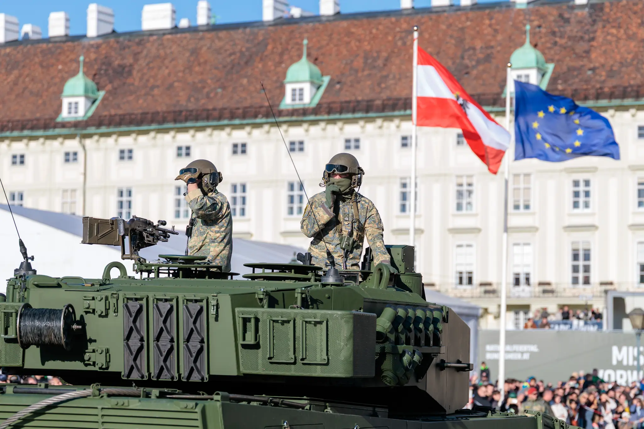 Zwei Soldaten in Tarnuniform stehen in der Luke eines Kampfpanzers vom Typ Leopard 2, dahinter wehen die österreichische und die europäische Flagge vor der Hofburg.