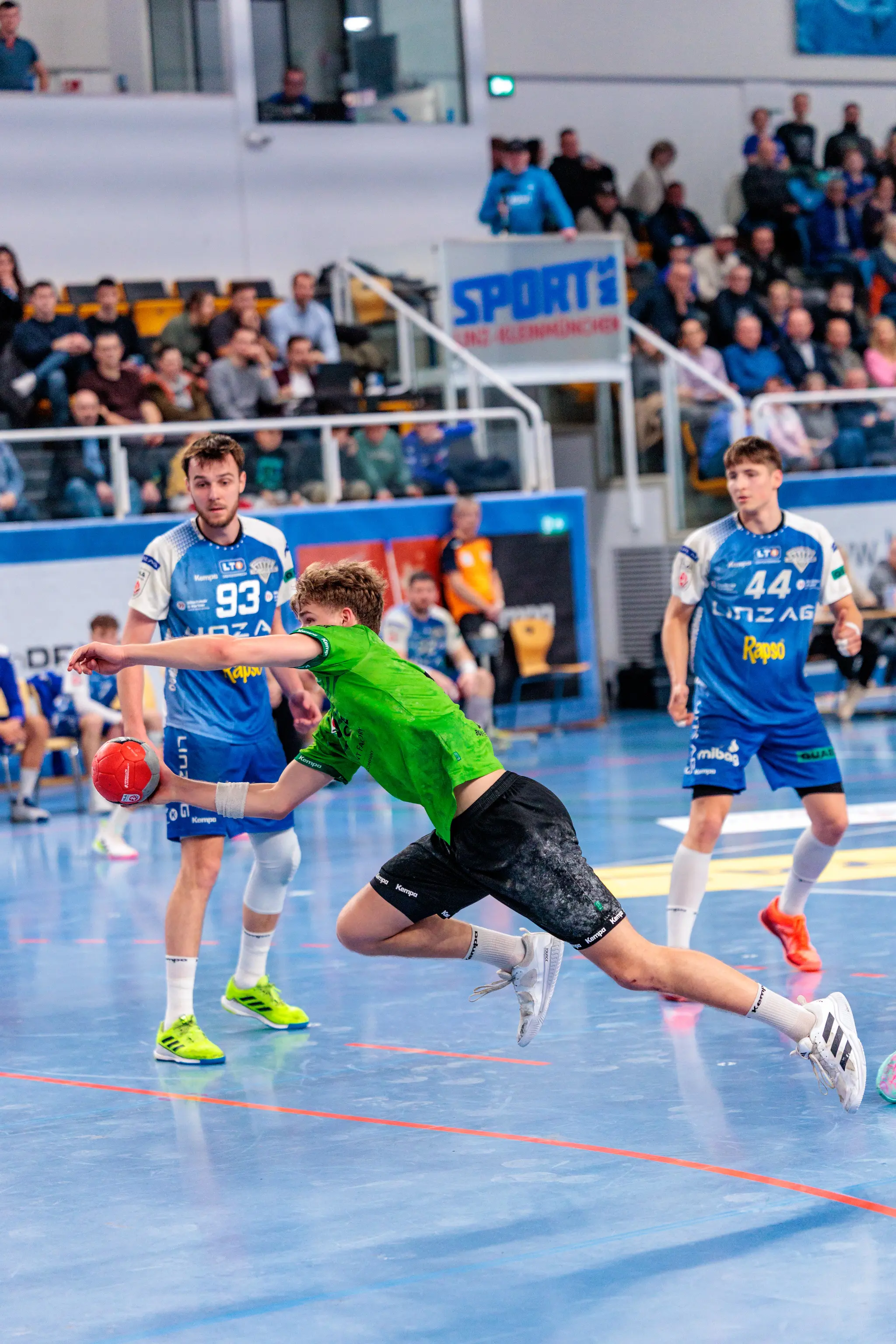 Handballspieler in grünem Trikot beim dynamischen Wurf auf blauem Spielfeld, umgeben von Zuschauern und Mitspielern 