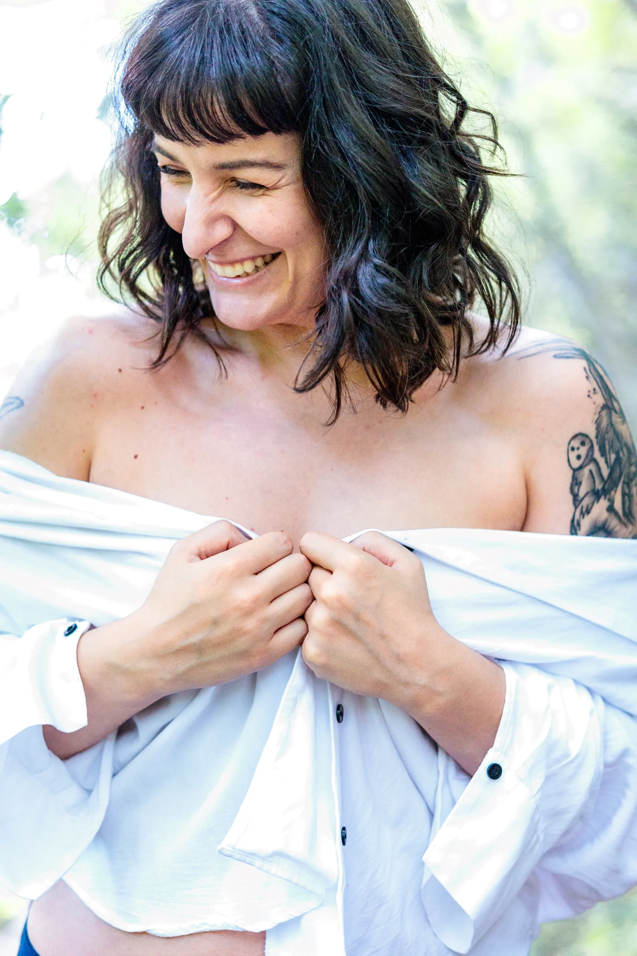 Eine lächelnde Frau mit dunklen Haaren und markanten Tattoos trägt ein schulterfreies, weißes Hemd vor einem weichen, grünen Hintergrund