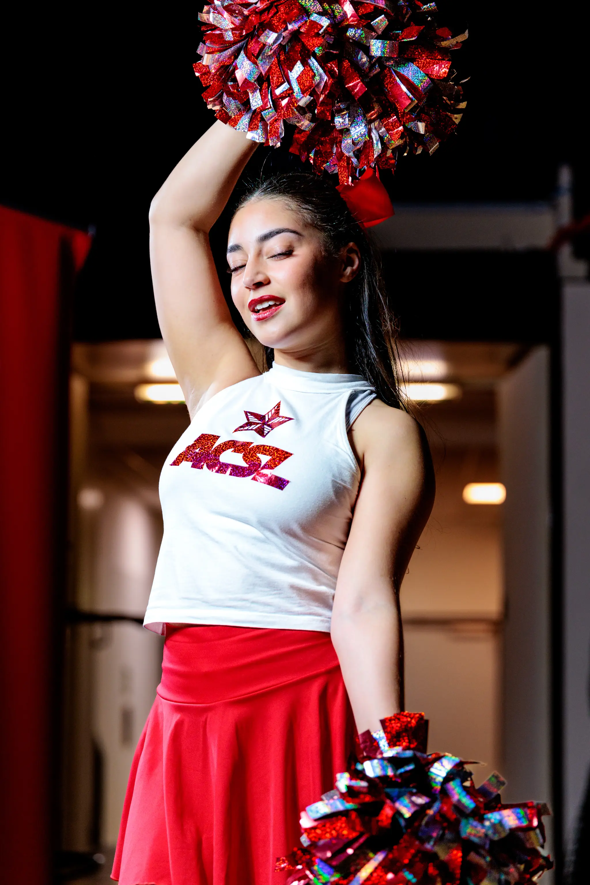 Eine junge Cheerleaderin in weißem ACSL-Top und rotem Rock hält glitzernde rote Pom-Poms in einer dynamischen Pose 