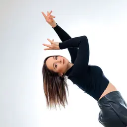 Eine junge Frau mit dunklem Haar und schwarzer Kleidung blickt in die Kamera, während sie in einer eleganten Tanzpose die Arme hebt