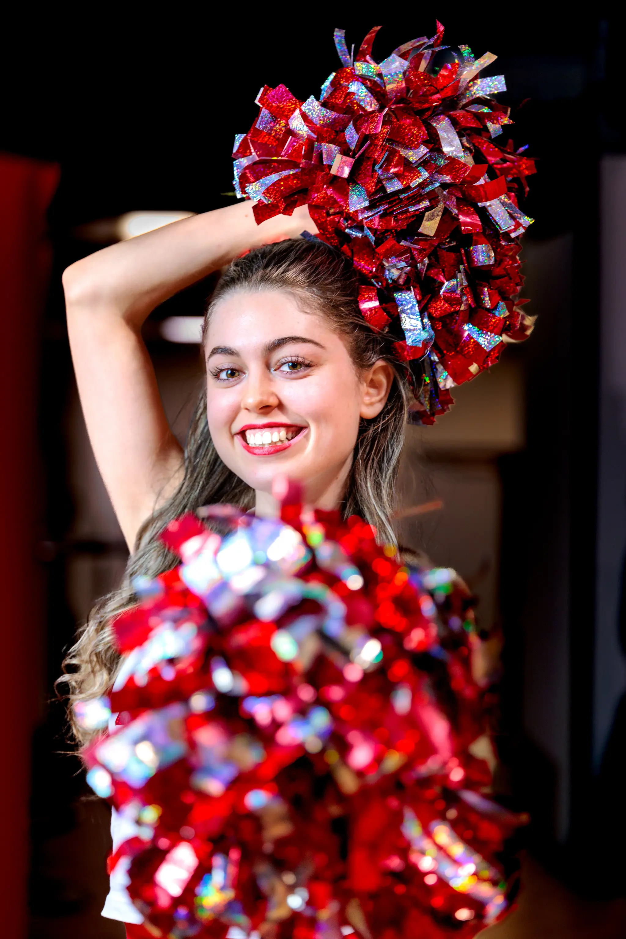 Eine junge Cheerleaderin in weißem ACSL-Top und rotem Rock hält glitzernde rote Pom-Poms in die Kamera 