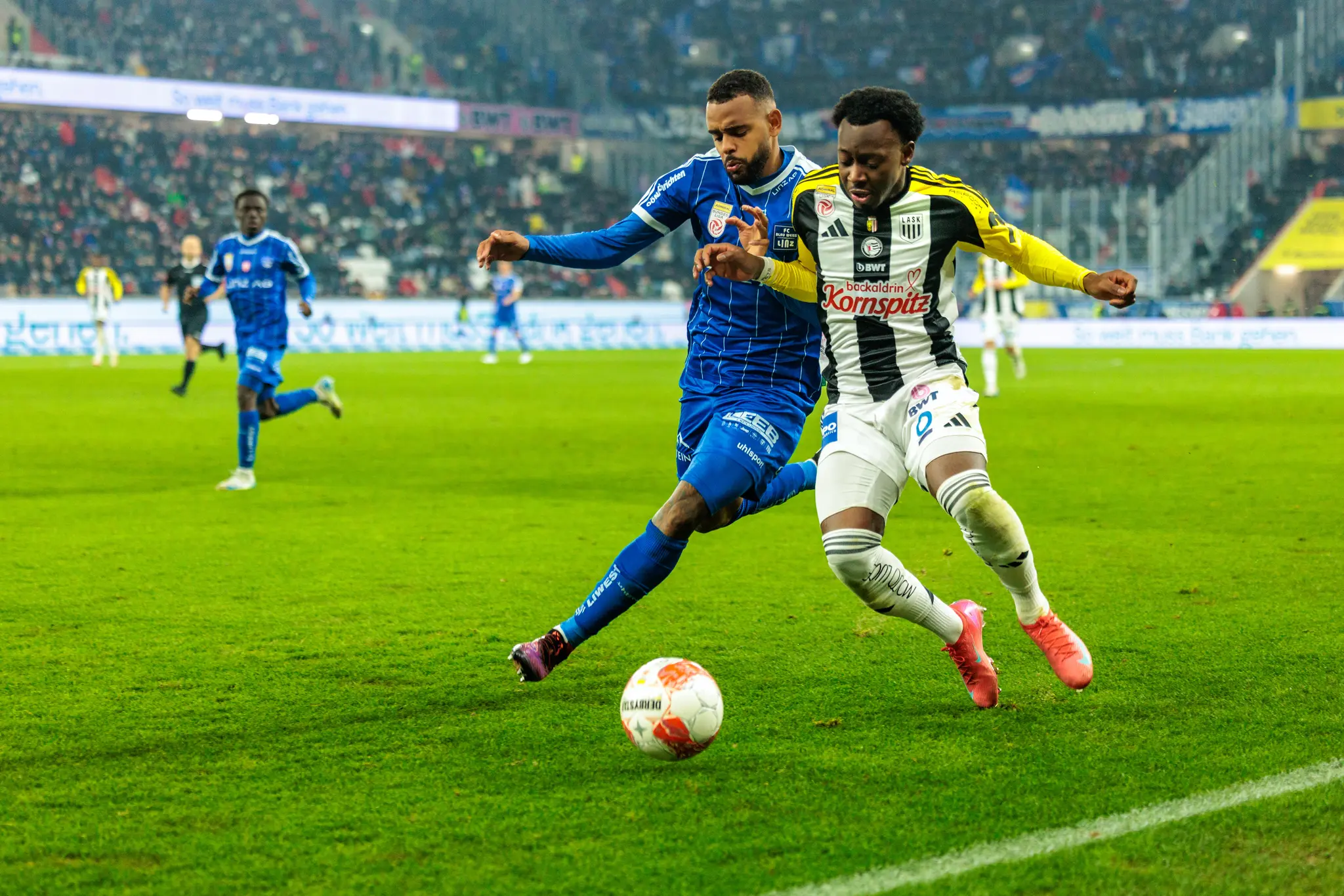 Zwei Fußballspieler, einer im blauen Trikot und einer vom LASK im gestreiften Trikot, sind im Zweikampf um den Ball auf dem Spielfeld der Raiffeisen Arena