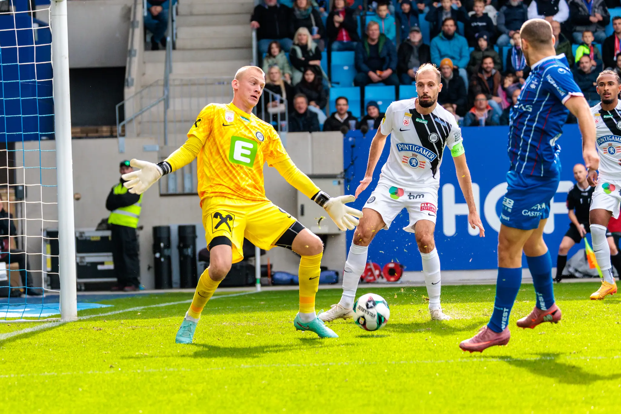 Sturm Graz-Torhüter Oliver Christensen in leuchtend gelber Tormannkleidung bei einer angespannten Abwehraktion im Strafraum gegen den FC Blau-Weiß Linz