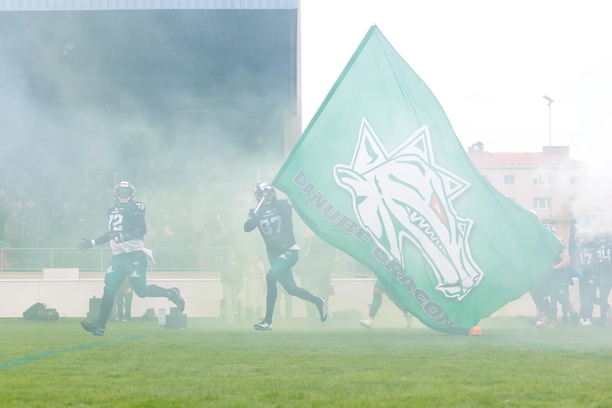 American-Football-Spieler der Danube Dragons laufen unter dichtem grünen Rauch und mit ihrer großen Teamflagge in ein Stadion ein