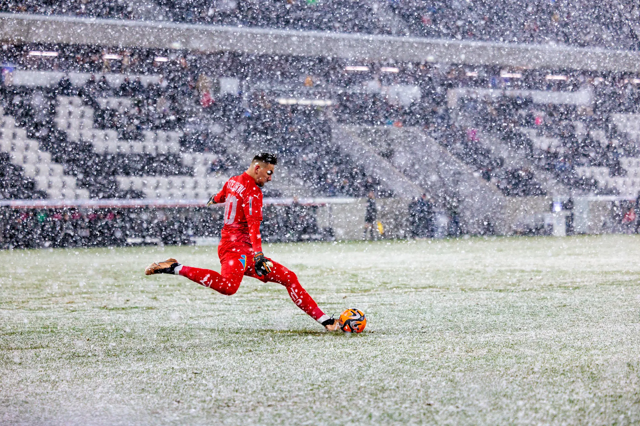 Ein Fußballtorwart im roten Trikot kickt einen orangefarbenen Ball auf dem schneebedeckten Feld eines Stadions während starken Schneefalls, mit Zuschauern auf den Tribünen im Hintergrund