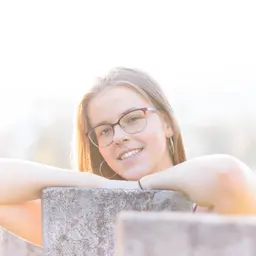 Eine junge Frau mit Brille lächelt und lehnt sich im goldenen Licht auf eine Betonmauer vor einem unscharfen Hintergrund lens: 56mm F1.4 DC DN | Contemporary 018
