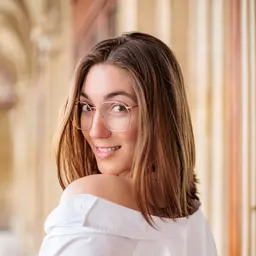 Eine lächelnde Frau mit Brille und weißem Hemd blickt über ihre Schulter direkt in die Kamera