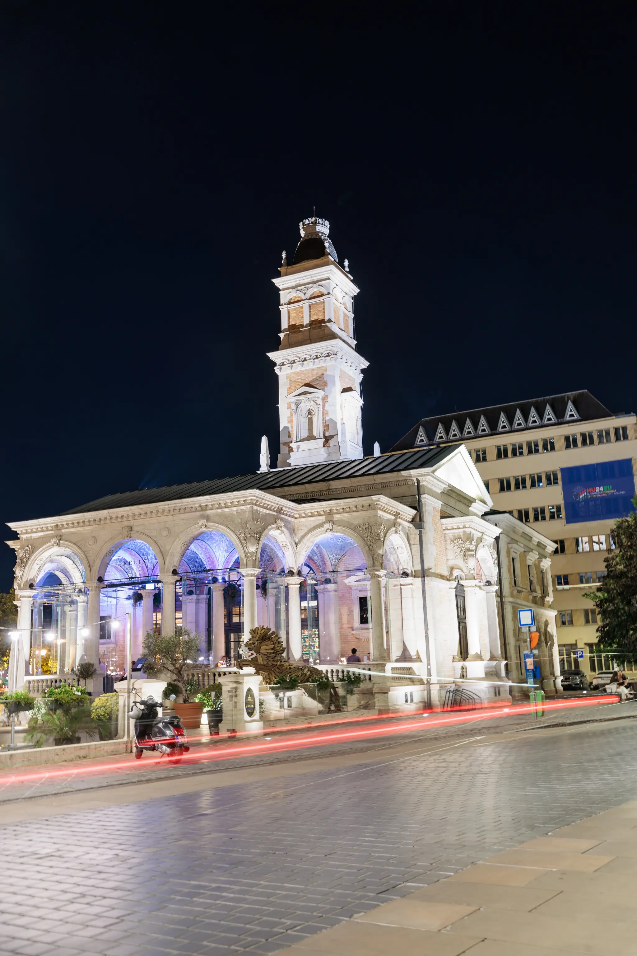 Nachtansicht eines imposanten, beleuchteten historischen Gebäudes mit Turm und Arkaden, davor eine Pflasterstraße mit roten Lichtspuren und einer Löwenskulptur. Eine urbane Nachtszene. 