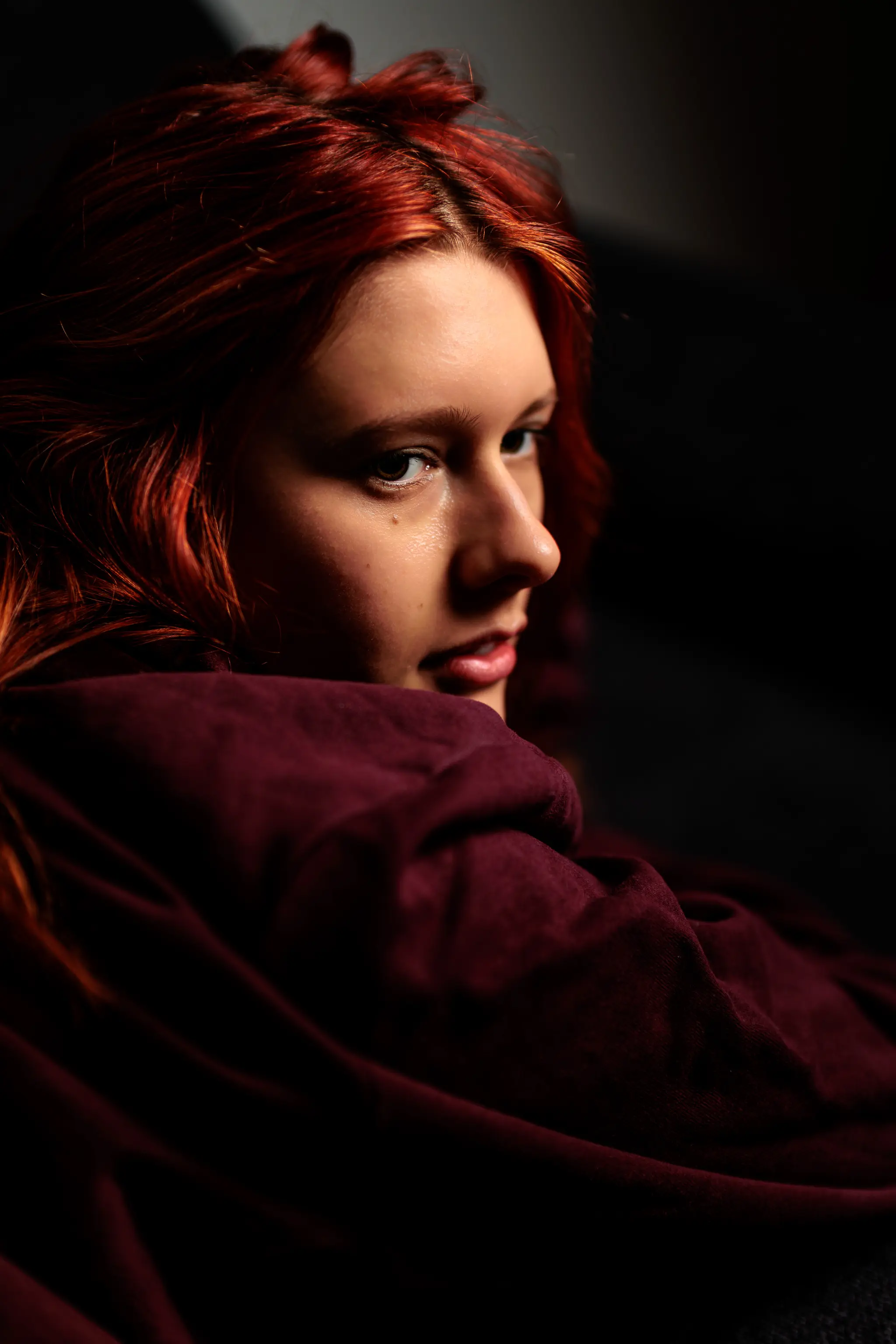 Nahaufnahme eines ausdrucksstarken Beauty-Portraits der rothaarigen Sarah in bordeauxrotem Hoodie, die mit intensivem Blick über die Schulter im stimmungsvollen Seitenlicht posiert