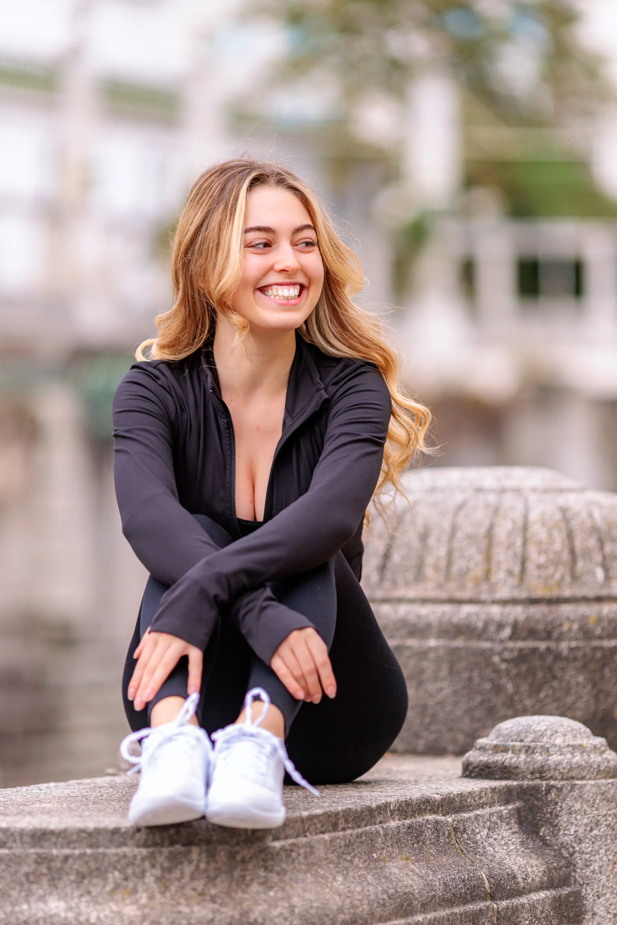 Model Valentina lacht herzlich, während sie in sportlicher schwarzer Kleidung auf einer Steinmauer im Wiener Stadtpark sitzt