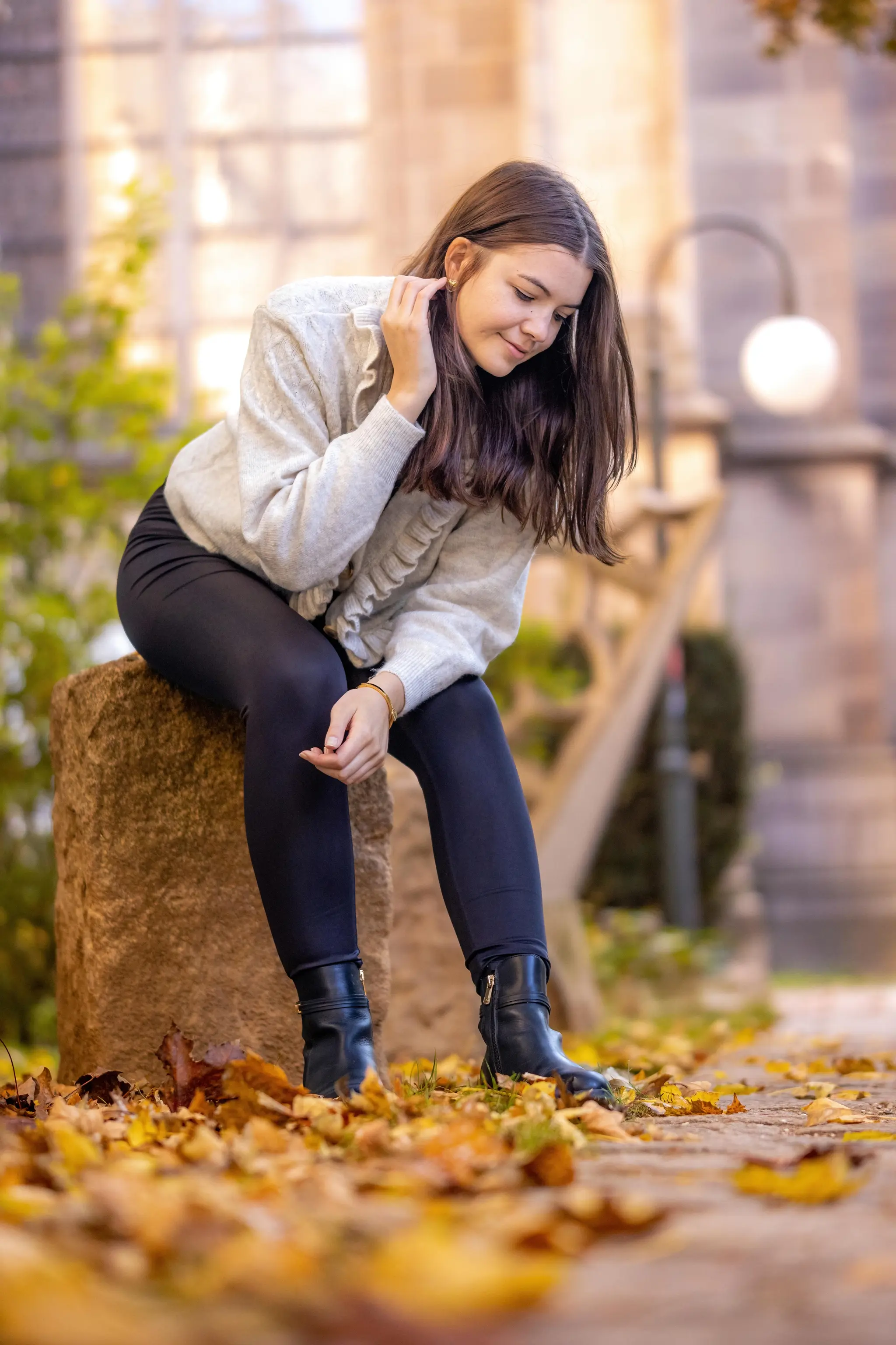 Eine junge Frau in einem hellen Pullover und dunklen Leggings sitzt auf einem Steinblock, umgeben von bunten Herbstblättern, und blickt lächelnd nach unten