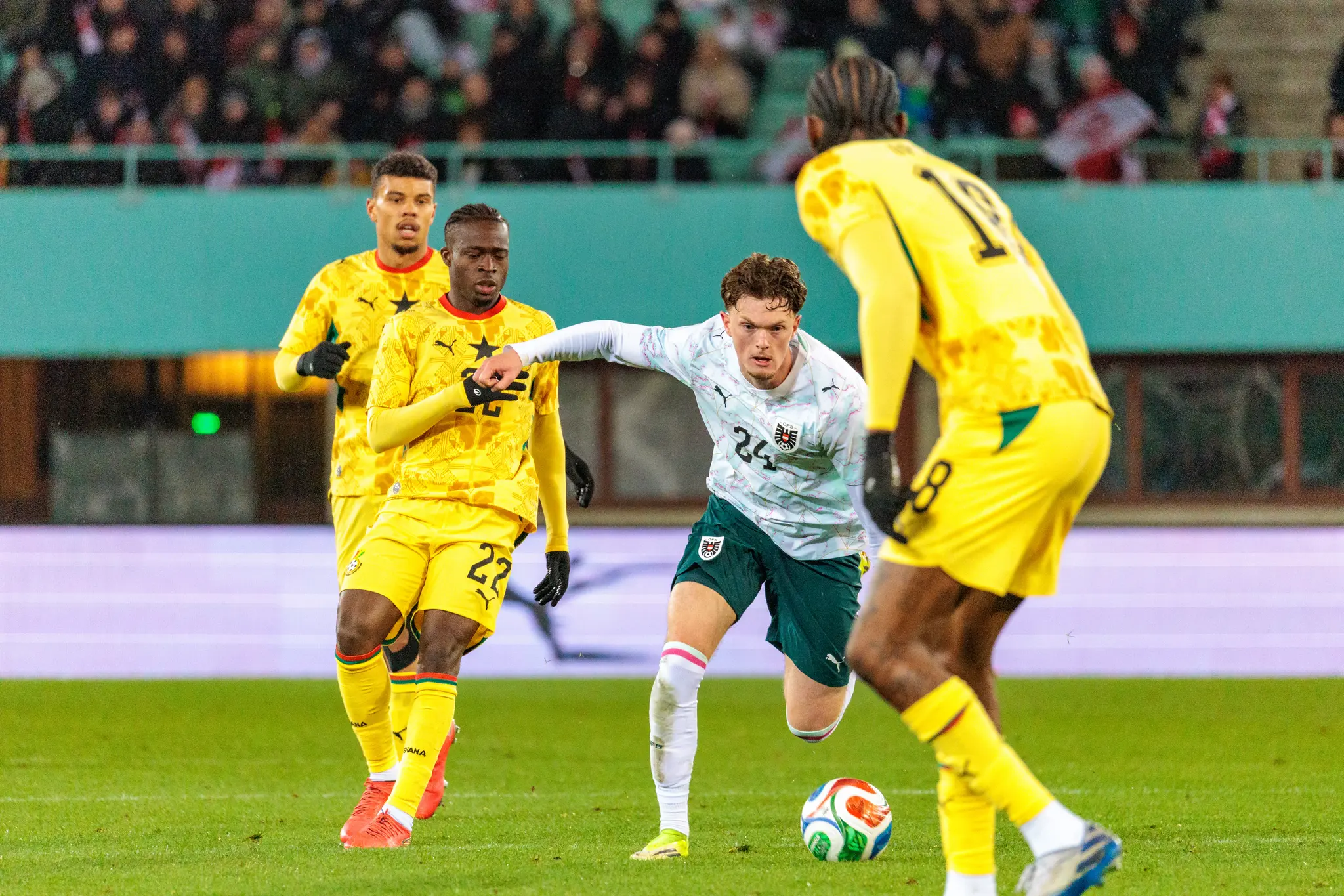 Ein österreichischer Spieler im weiß-grünen Trikot im intensiven Zweikampf um den Fußball mit einem ghanaischen Gegenspieler im gelben Trikot, begleitet von weiteren Ghana-Spielern
