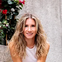 Nahaufnahme einer lächelnden Frau mit langen blonden Locken und Perlenkette vor einem leicht unscharfen Hintergrund aus einer strukturierten Wand und Pflanzen