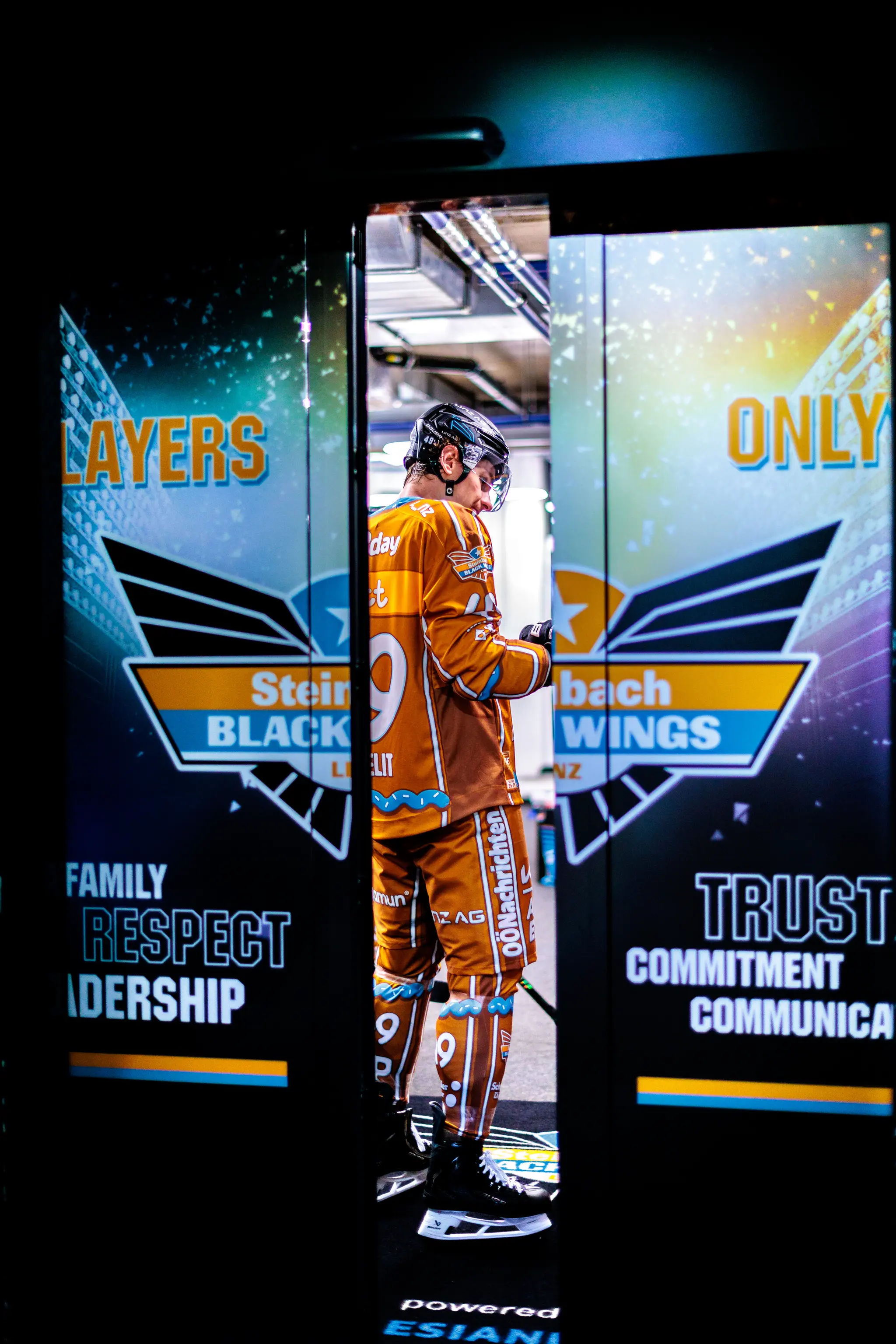 Eishockeyspieler Graham Knott der Steinbach Black Wings Linz in orangem Trikot mit der Nummer 49 steht nachdenklich in einem beleuchteten Spielertunnel mit Team-Logos