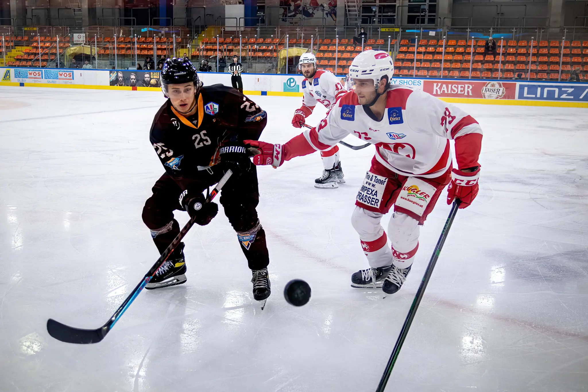 Ein Eishockeyspieler der SteelWings Linz im schwarzen Trikot führt den Puck auf dem Eis, während ein Gegenspieler im rot-weißen Trikot ihn bedrängt