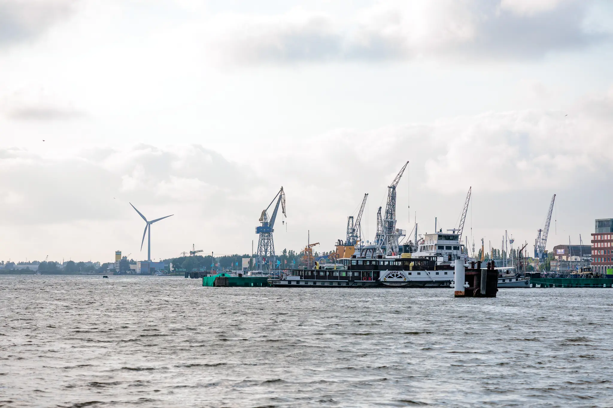 Blick über das Wasser auf den Amsterdamer Hafen mit Schiffen, Kränen und einer Windturbine unter Wolken 