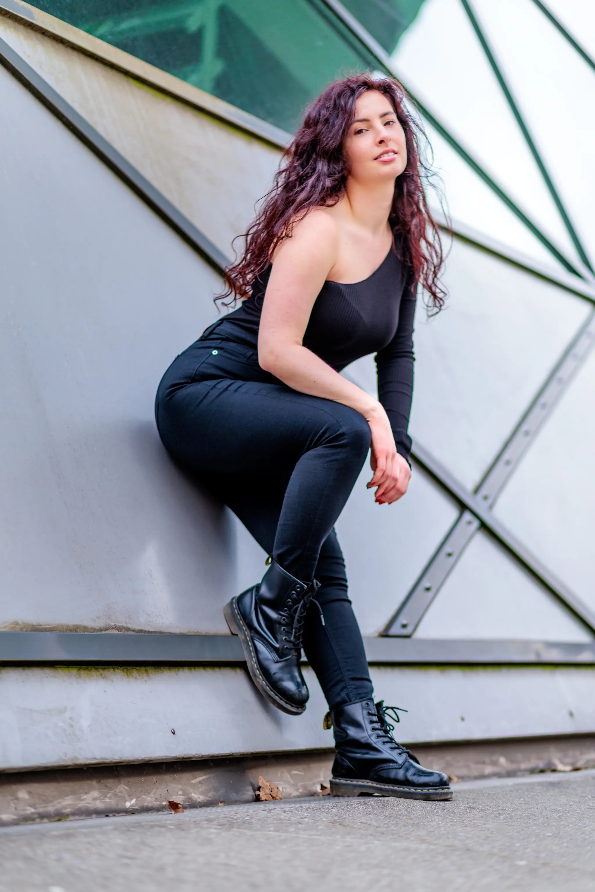 Junge Frau mit dunklen Locken in schwarzem One-Shoulder-Top, schwarzer Jeans und schwarzen Stiefeln, lässig an einer grauen Wand gelehnt, Outdoor-Porträt
