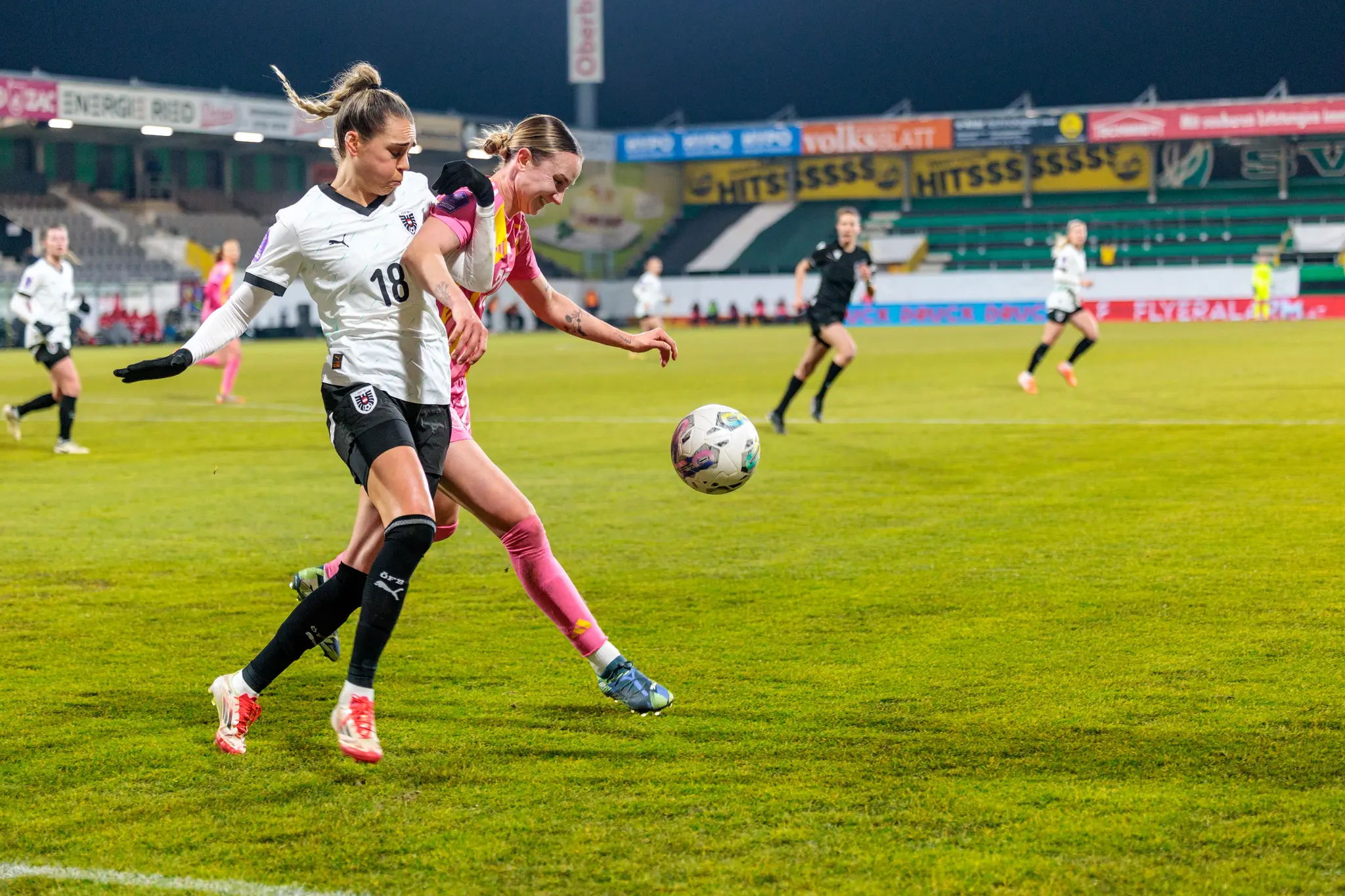 Eine österreichische Fußballerin in Weiß mit der Nummer 18 und eine Gegnerin in Pink kämpfen während eines Spiels dynamisch um den hoch springenden Ball