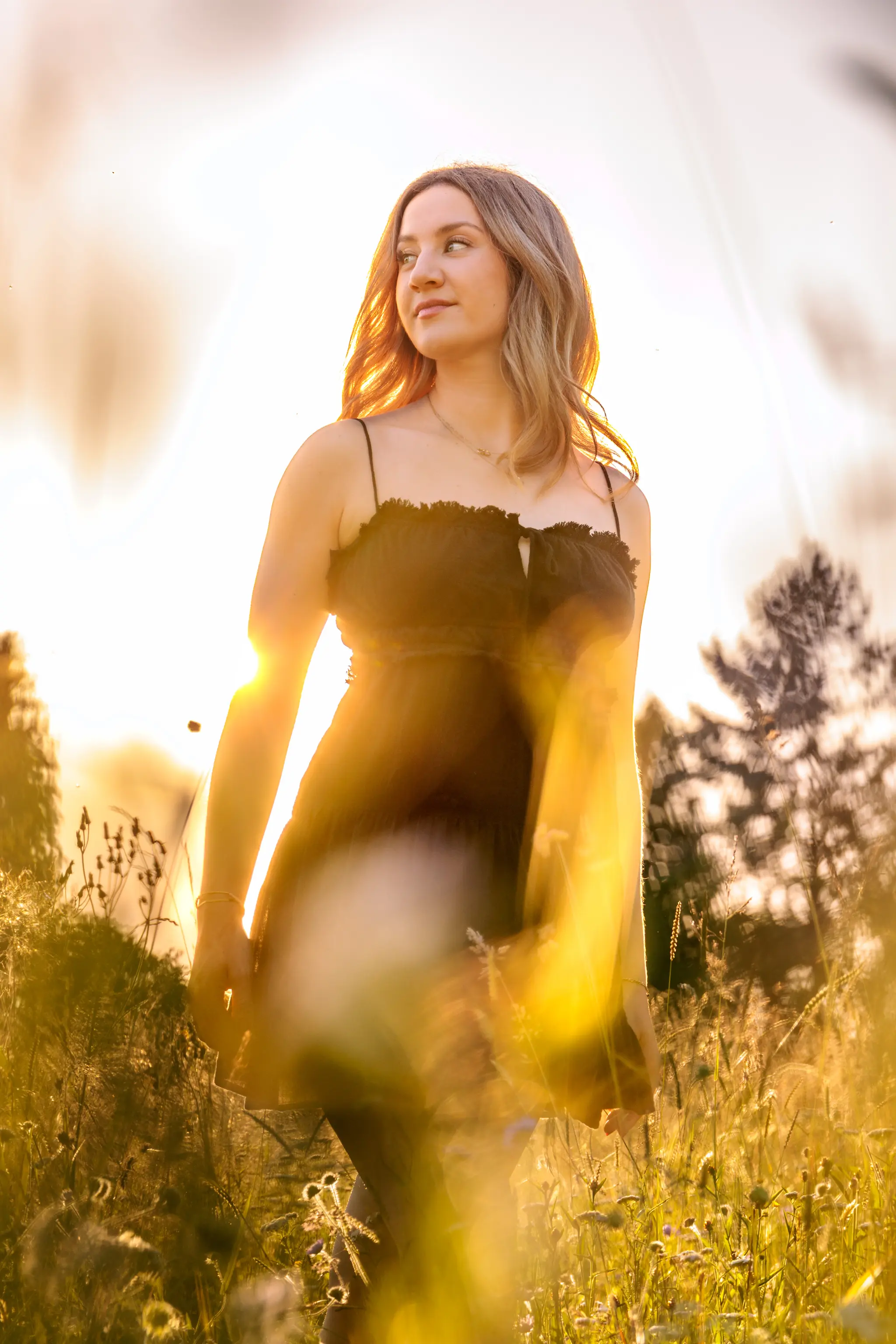Eine junge Frau im dunklen Kleid steht bei goldenem Gegenlicht in einer sonnigen Sommerwiese 