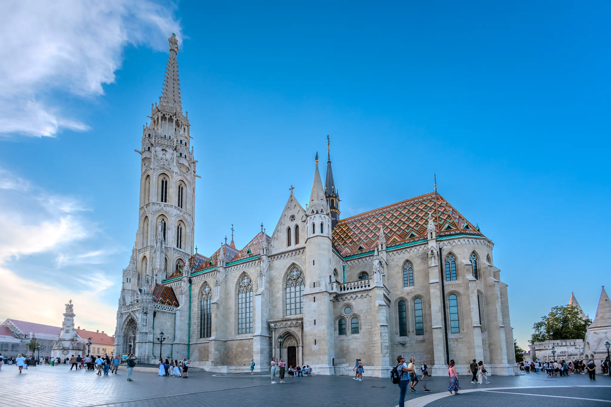 Die Matthiaskirche in Budapest, Ungarn, ein historisches Gebäude mit einem markanten bunten Ziegeldach und hohem Turm, umgeben von Touristen unter blauem Himmel. 