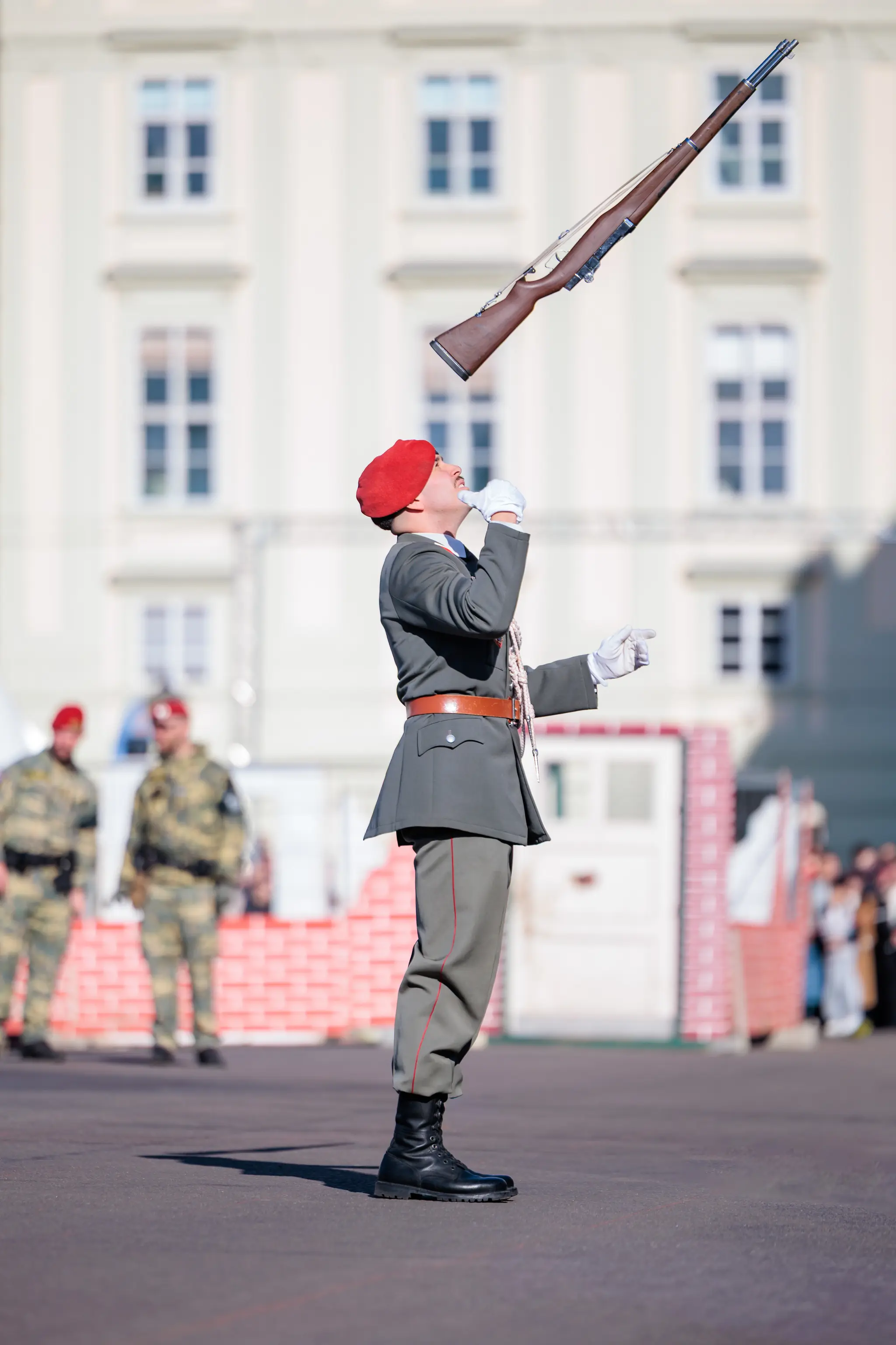 Ein Gardesoldat mit rotem Baret wirft ein Gewehr hoch in die Luft, den Kopf in den Nacken gelegt, hinter ihm ein helles Gebäude.