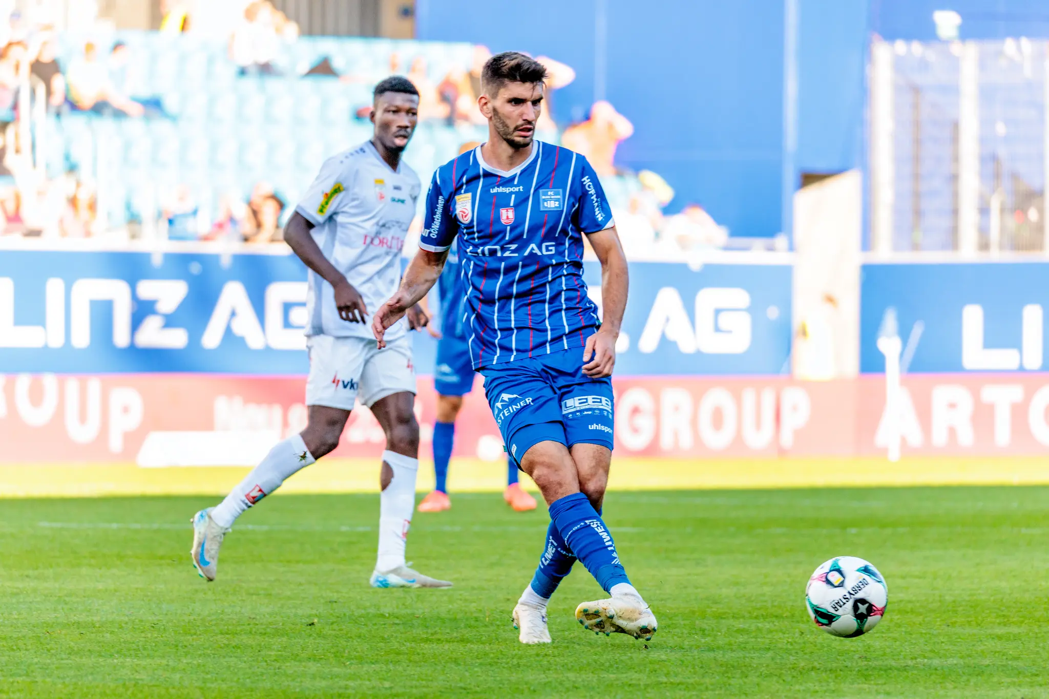 Der Mittelfeldspieler des FC Blau-Weiß Linz, Christopher Cvetko, kontrolliert in einer dynamischen Spielszene den Fußball, während ihn ein Gegenspieler im Hintergrund beobachtet