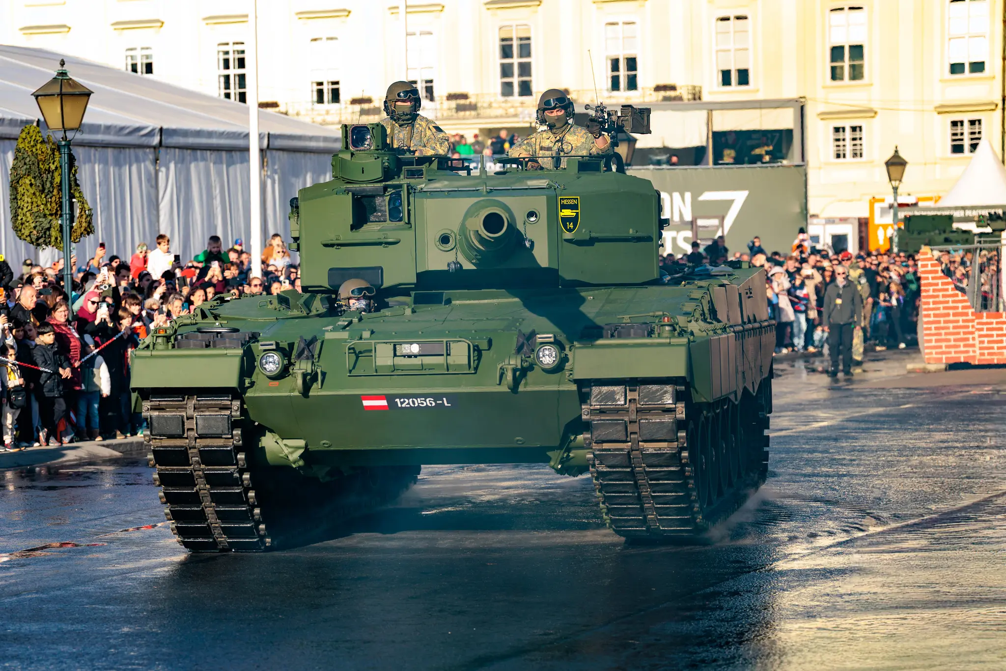 Ein Kampfpanzer vom Typ Leopard 2 rollt über den nassen Heldenplatz, am linken Rand verfolgt das Publikum die Fahrt.