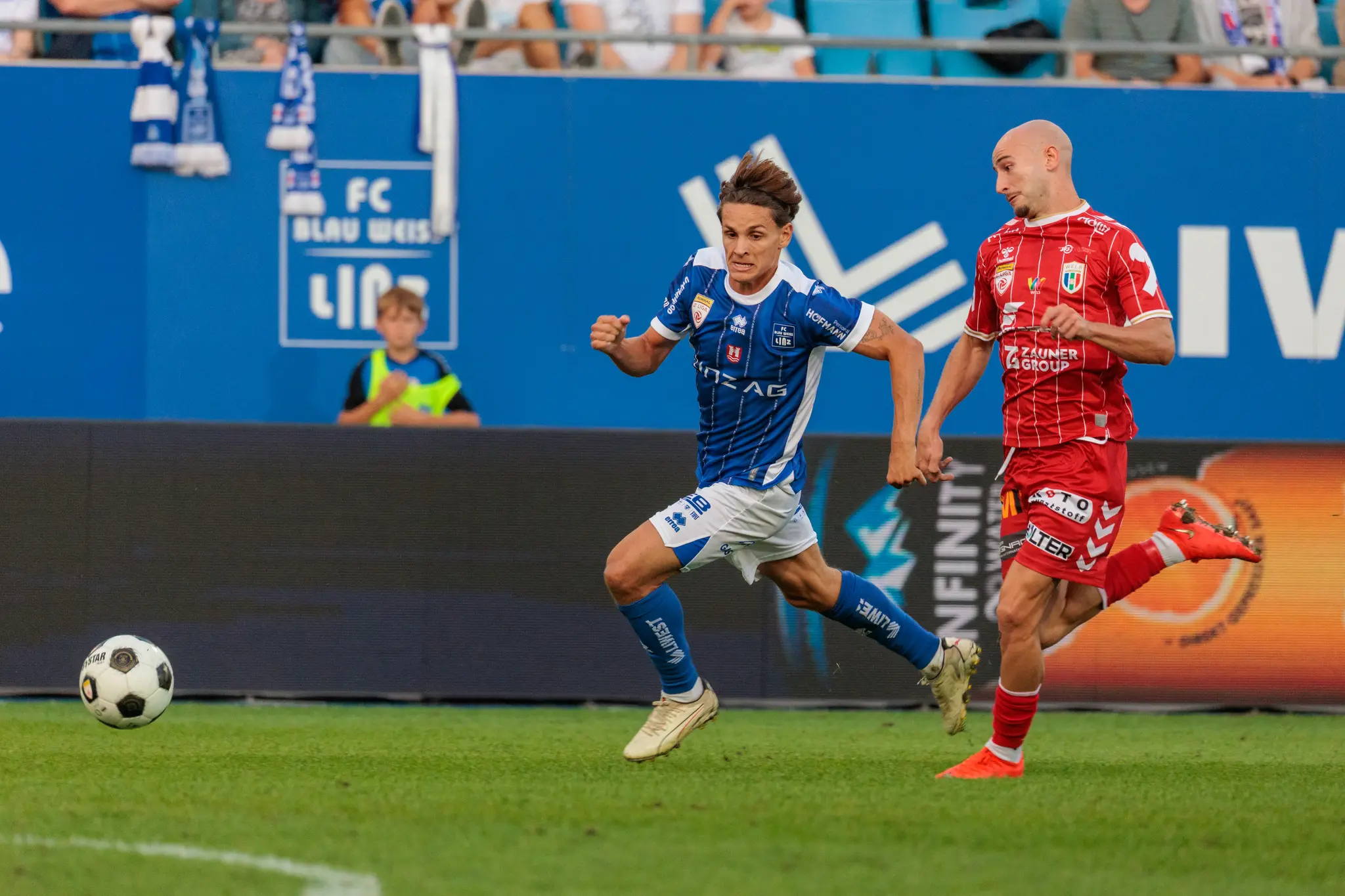 Ein Linzer Spieler sprintet mit angezogenem Arm dem Ball entgegen, dicht gefolgt von einem Welser Spieler; die Bande mit dem Blau-Weiß-Logo säumt die Szene.
