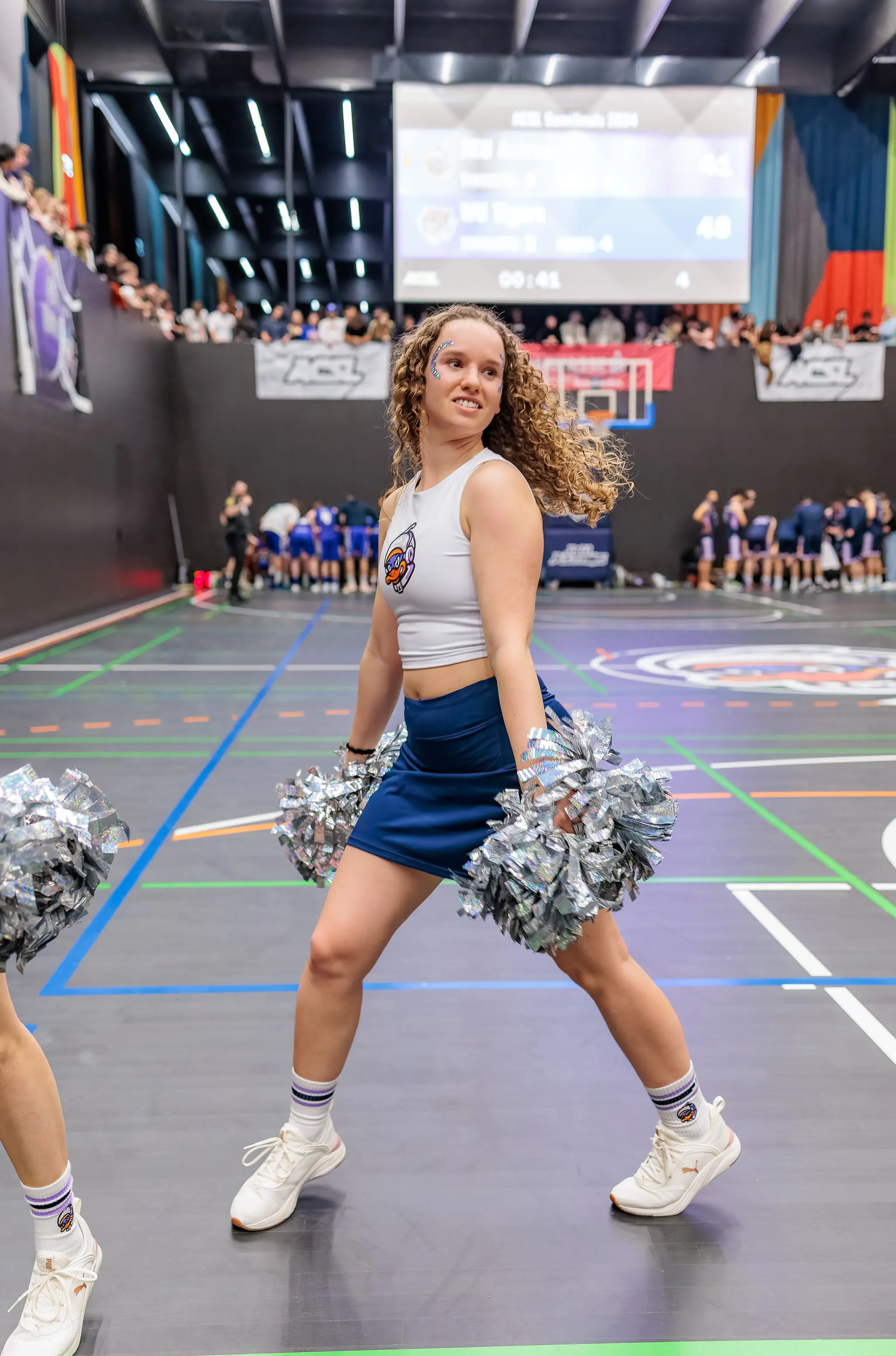 Eine lächelnde Cheerleaderin in Weiß und Blau hält Pom-Poms während eines ACSL Basketball-Halbfinales 2024 