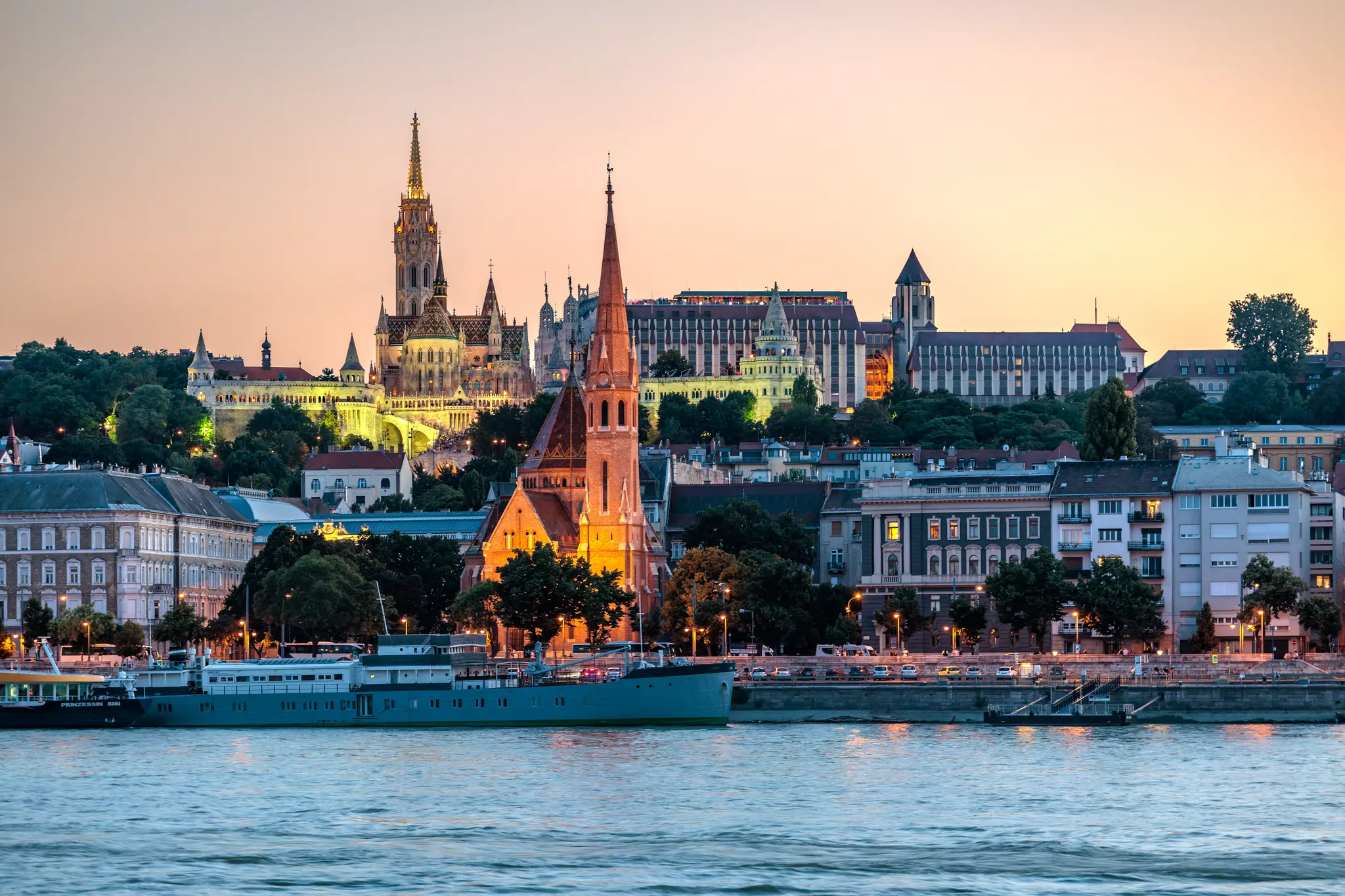Stadtansicht von Budapest bei Sonnenuntergang mit der beleuchteten Matthiaskirche, Fischerbastei und dem Budaer Burgviertel am Donauufer.
