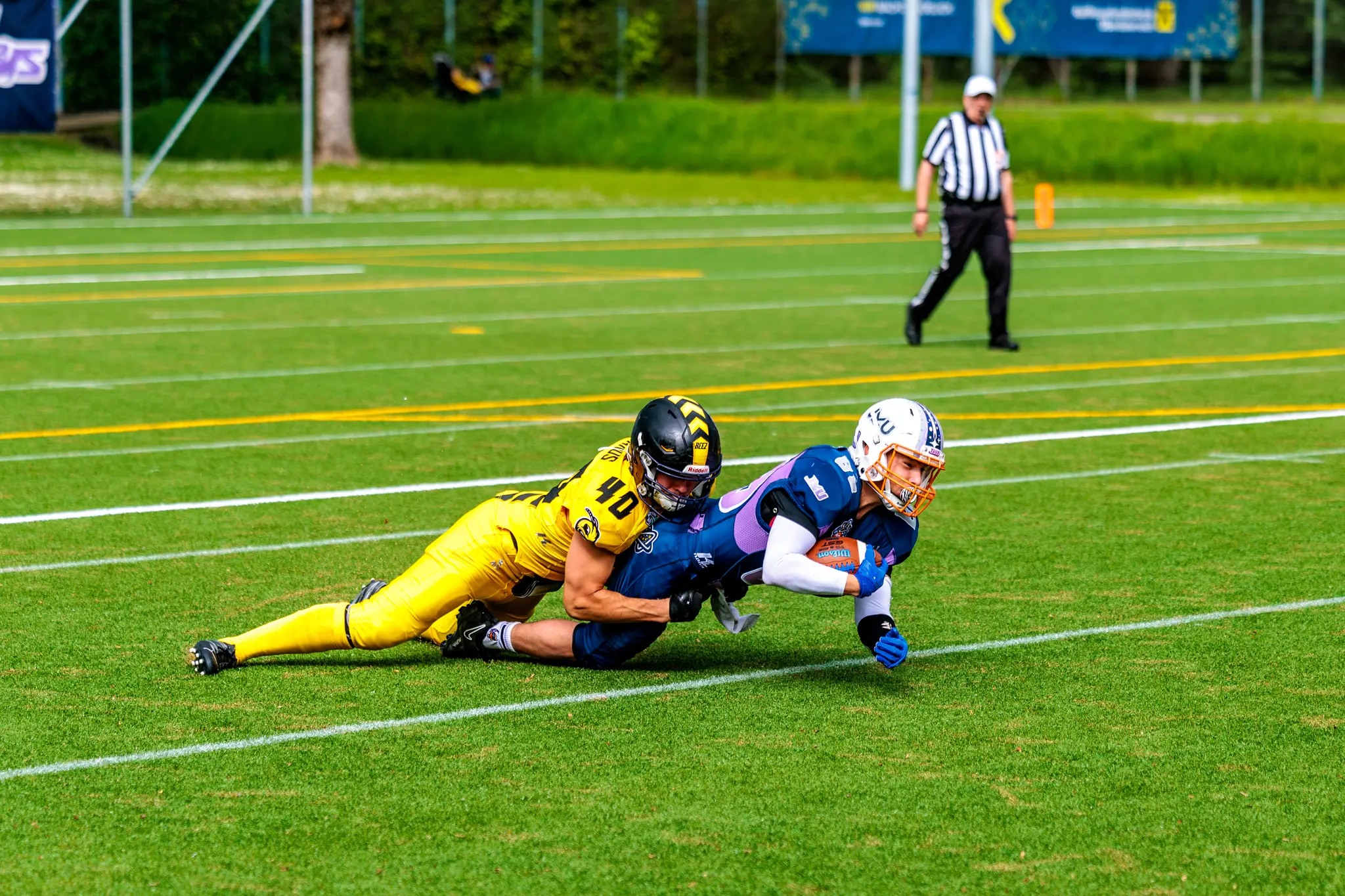 Ein American-Football-Spieler im lila Trikot versucht, einen Touchdown zu erzielen, während er von einem Spieler im gelben Trikot getackelt wird