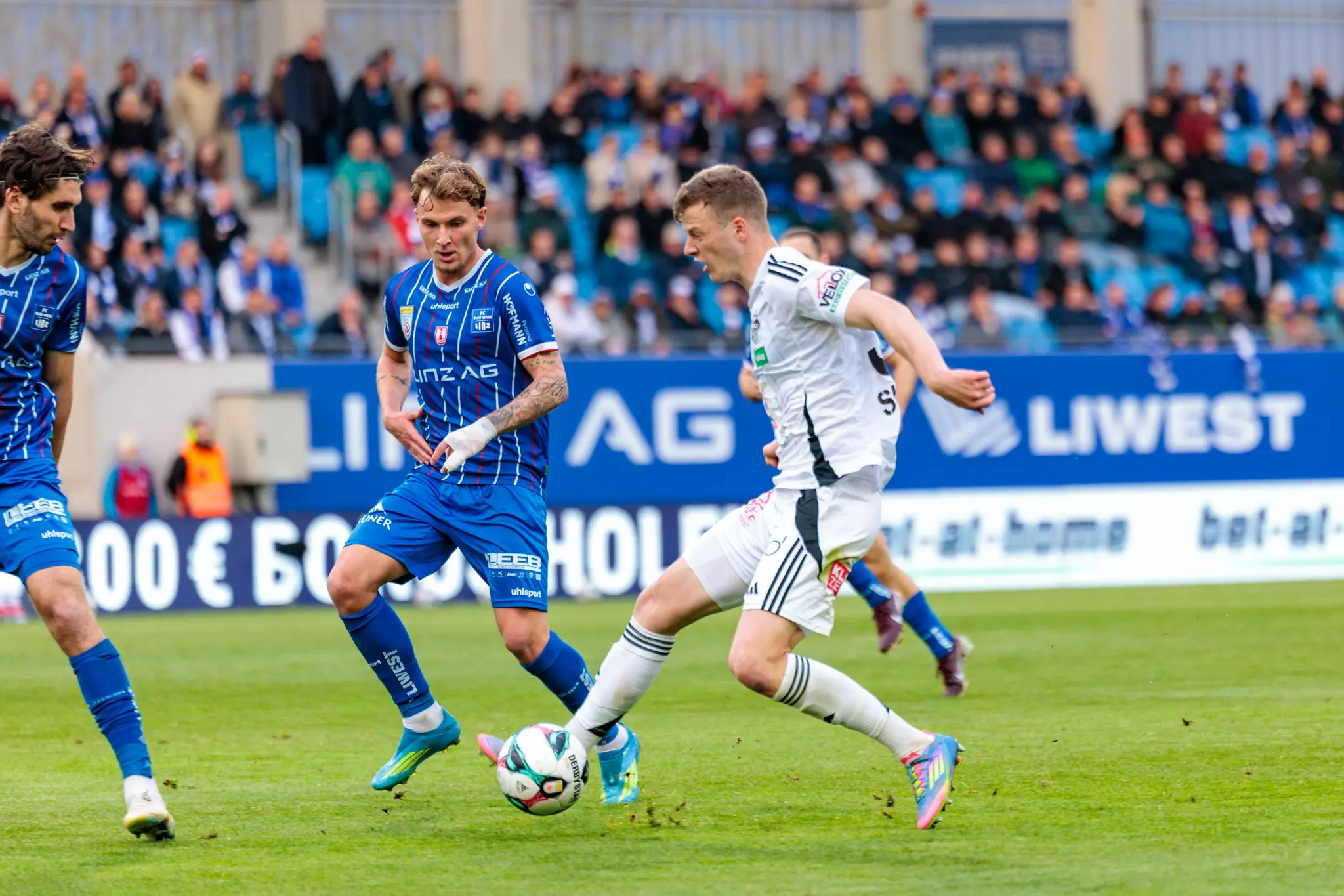 Ein Spieler des Wolfsberger AC kämpft in einem intensiven Zweikampf um den Ball gegen zwei Gegenspieler des FC Blau-Weiß Linz