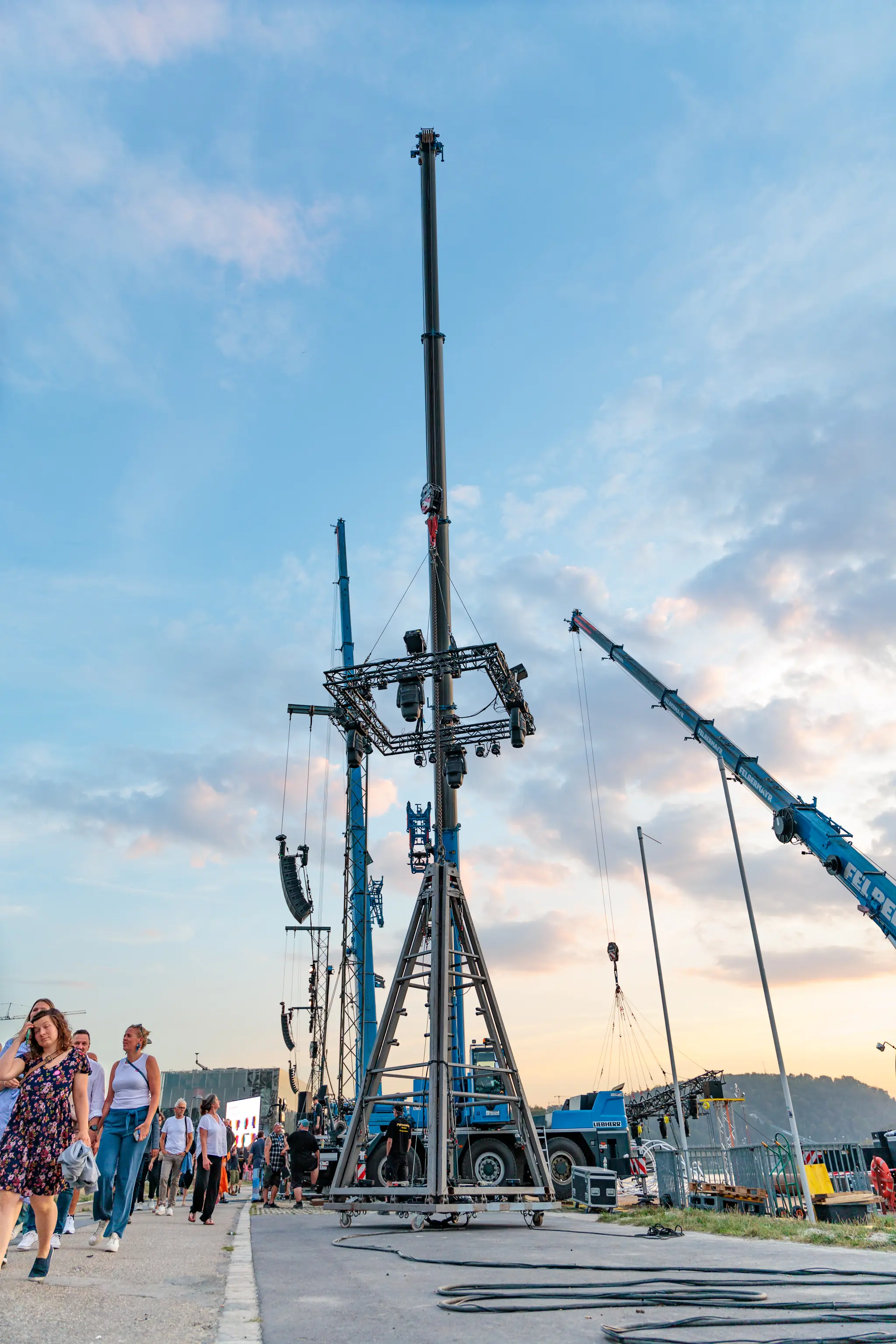 Konzertbühnenaufbau im Freien mit hohen Kränen und Rigg-Türmen für Licht- und Tontechnik. Besucher und Personal auf dem Veranstaltungsgelände unter bewölktem Himmel 