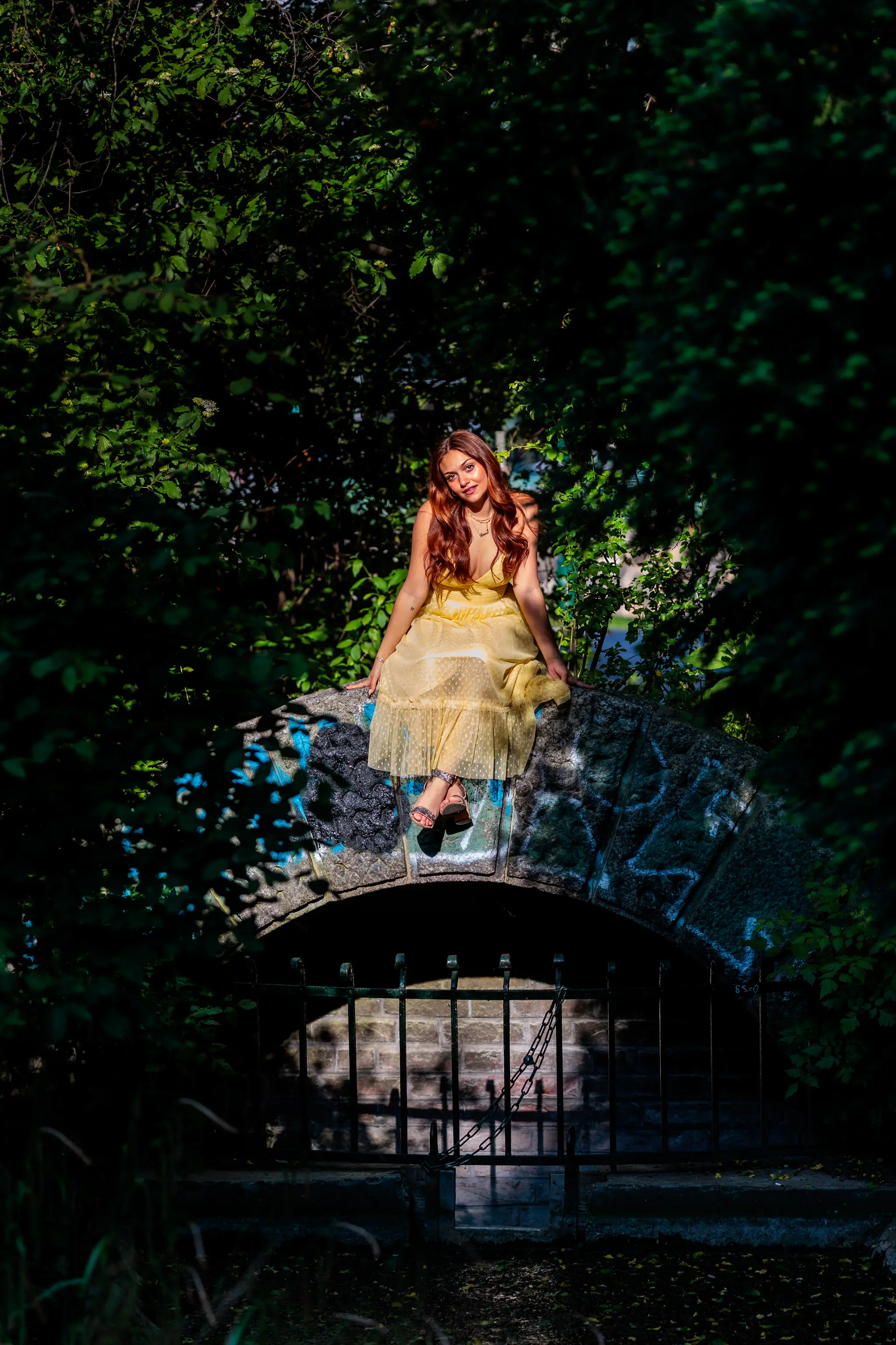 Junge Frau im gelben Sommerkleid sitzt im Sonnenlicht auf einer alten Steinbogenbrücke mit Graffiti, umrahmt von dichtem Grün. 