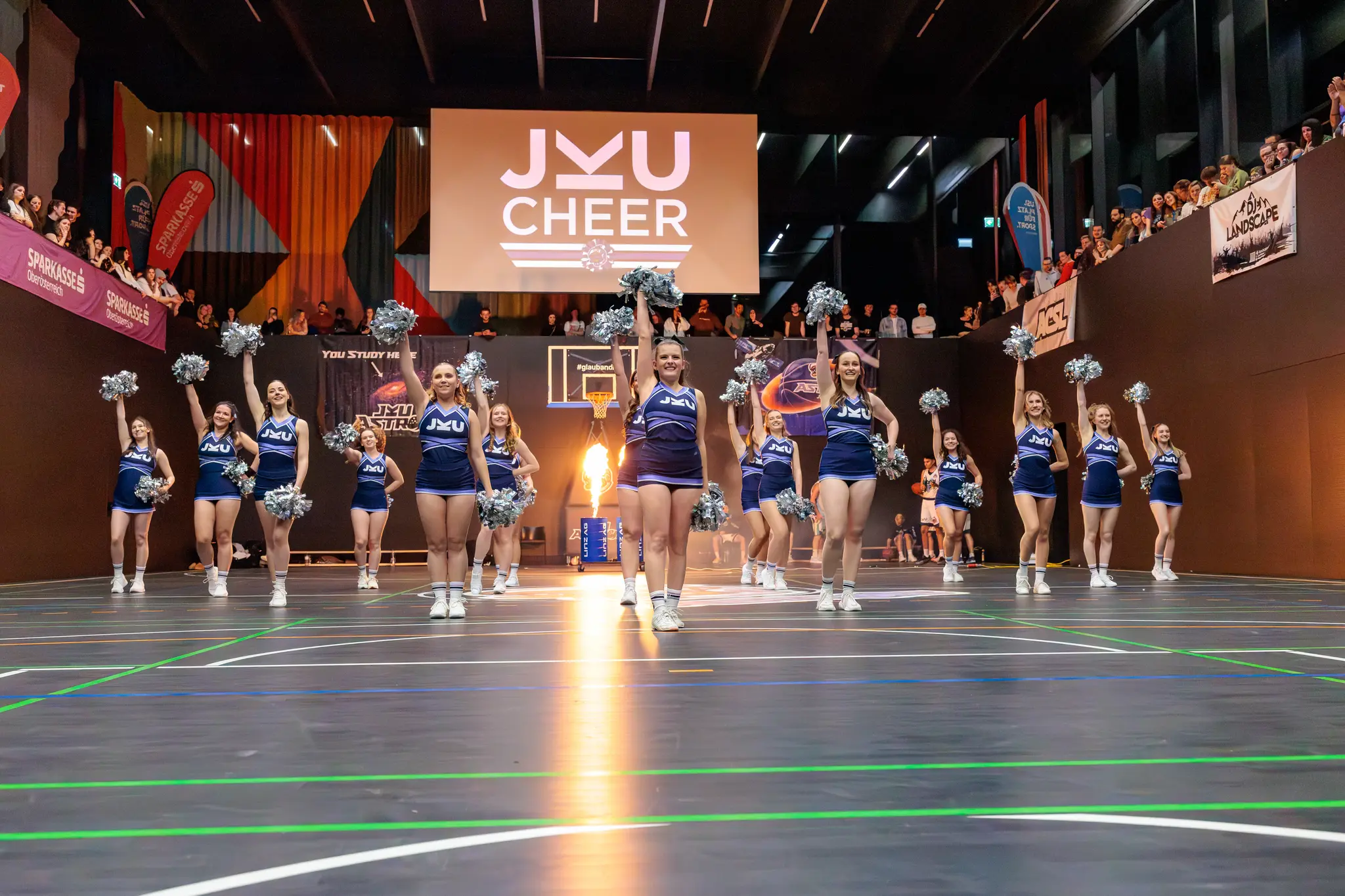 JKU CHEER Cheerleader-Team in blauen Uniformen performt mit Pom-Poms in einer Sporthalle vor Publikum und großem Logo-Bildschirm 