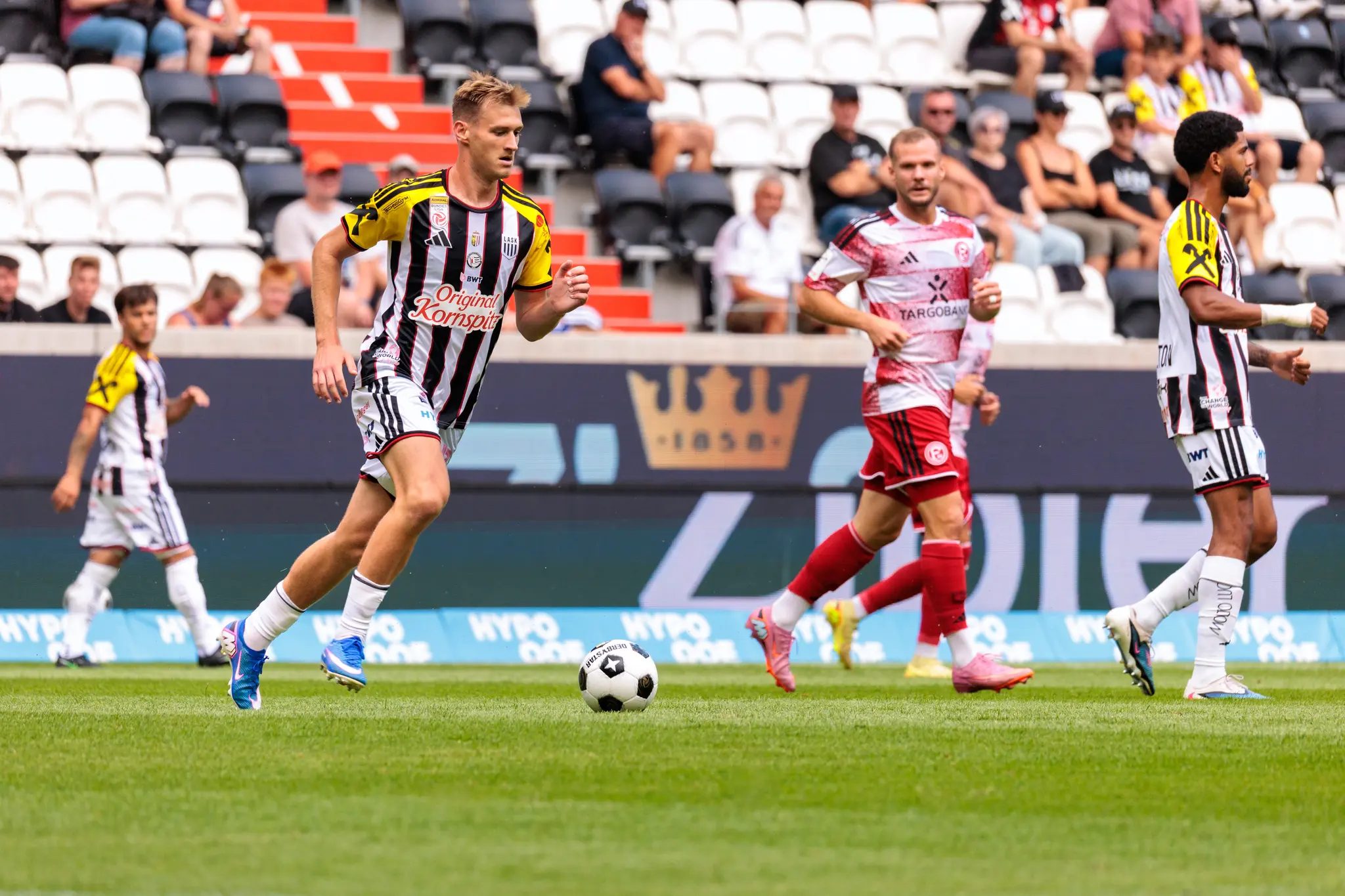 Junger LASK-Spieler mit blonden Haaren kontrolliert den Ball, während ein Düsseldorfer Spieler und weitere Teamkollegen im Hintergrund positioniert sind.