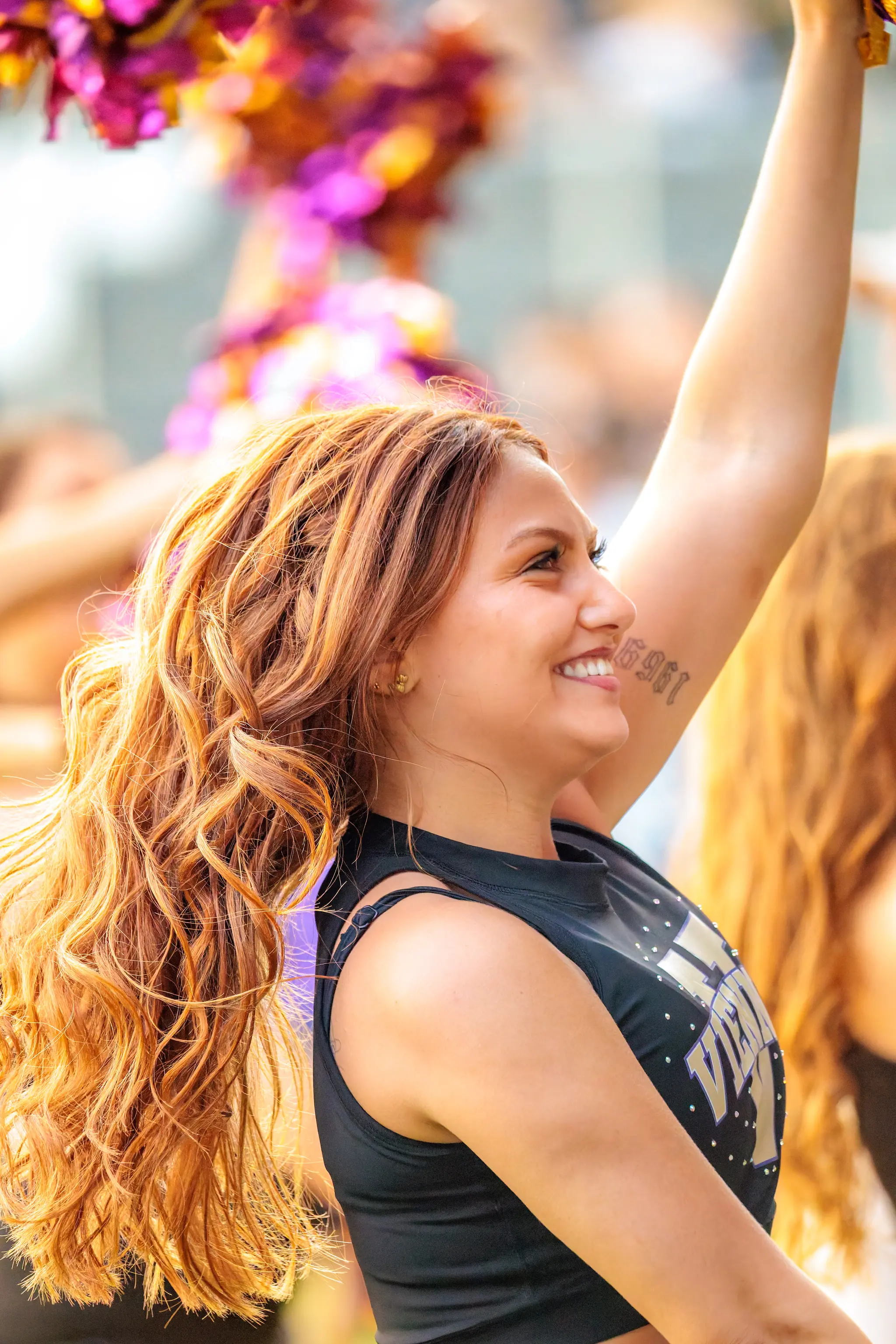 Profil-Portrait einer Tänzerin des Vienna Vikings Cheer Squads kurz vor dem Anpfiff: lächelnd, den Pompon in der erhobenen Hand, im warmen Abendlicht des Wiener Sport-Club-Stadions.