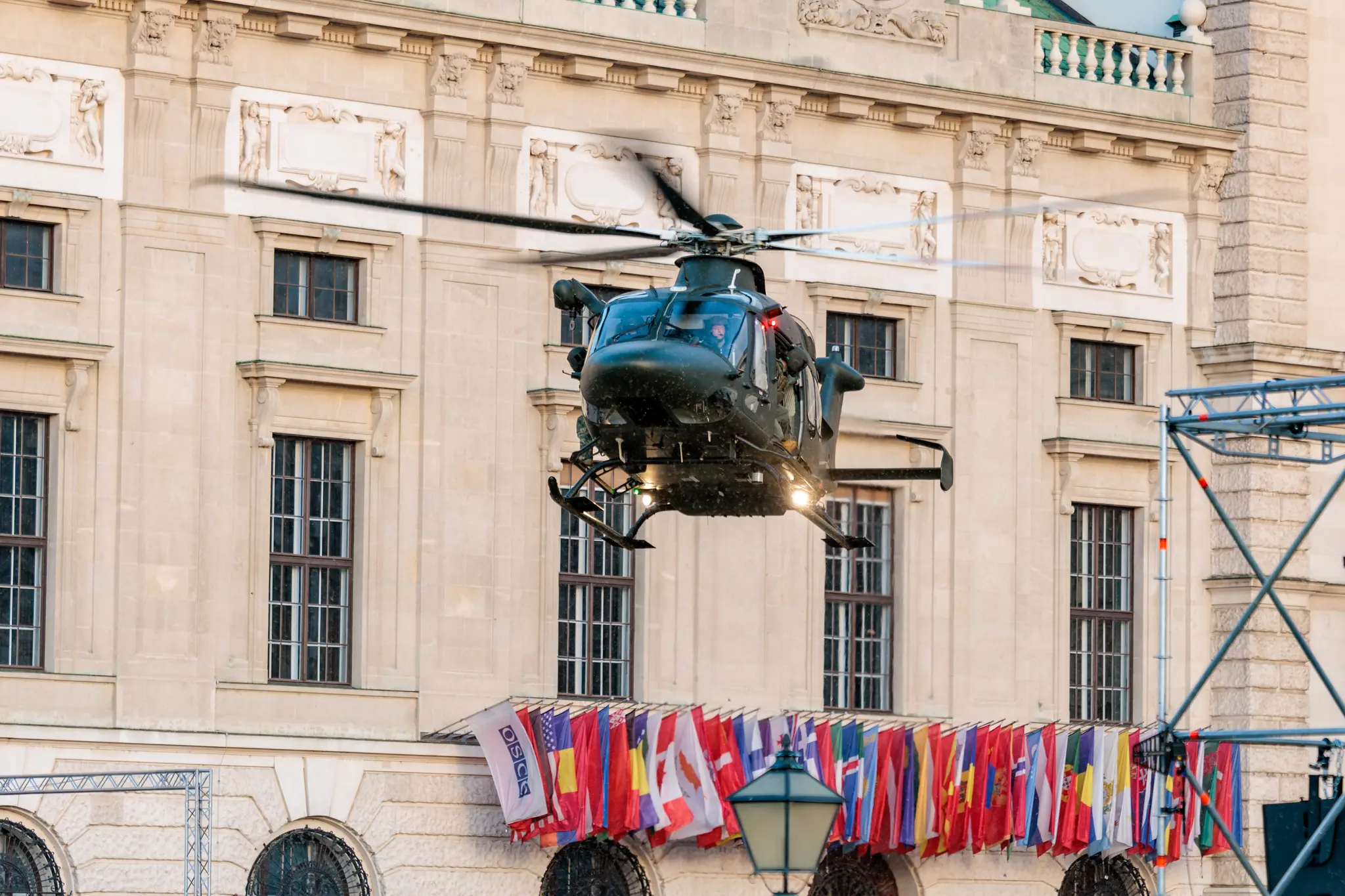Ein Militärhubschrauber vom Typ Leonardo AW169M „Lion“ schwebt vor der Fassade der Hofburg, darunter säumen zahlreiche internationale Flaggen den Balkon.